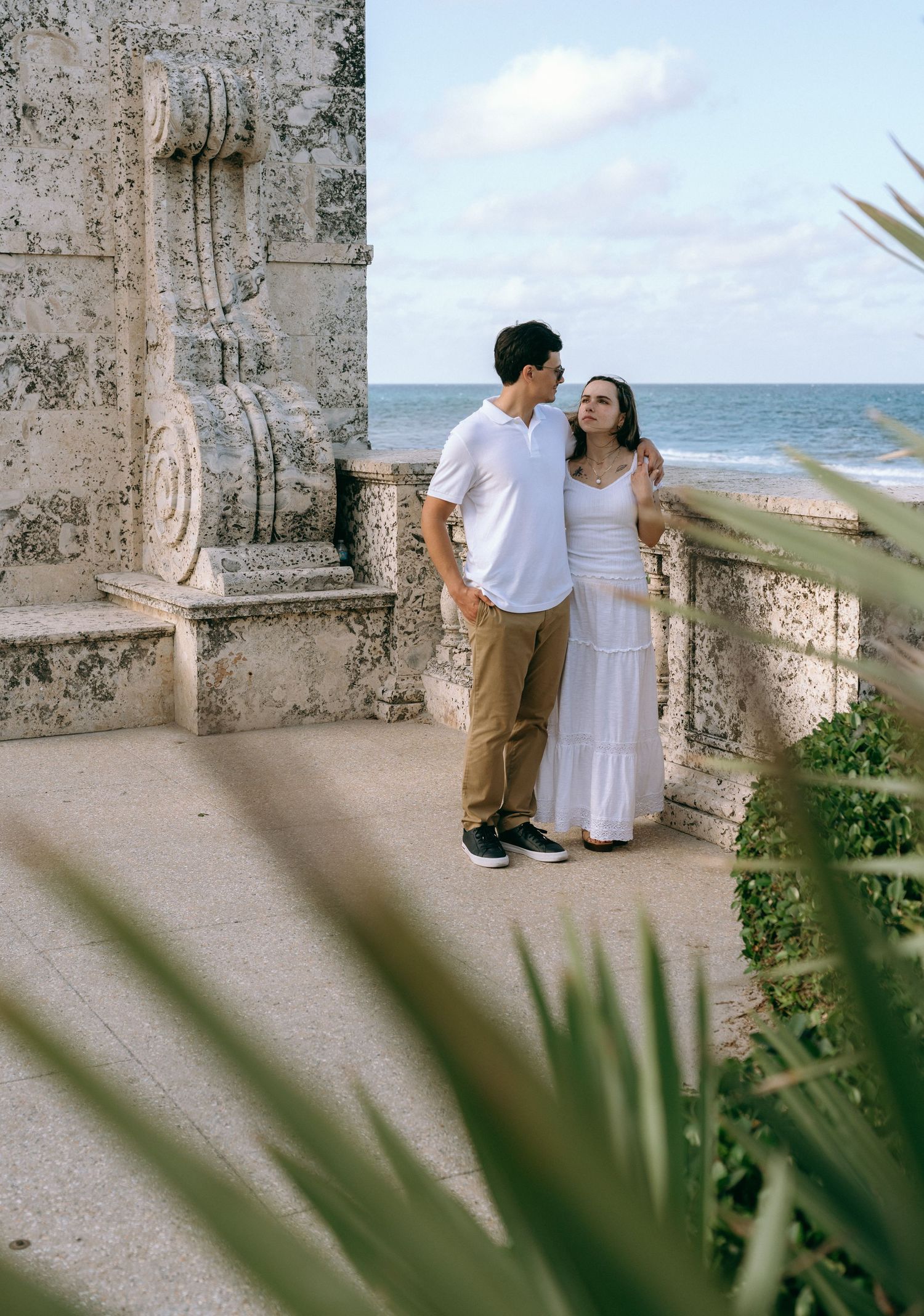 Couple in white outfits standing together on a stone terrace overlooking the ocean. Engagement photos on Worth Avenue in Palm Beach, Florida