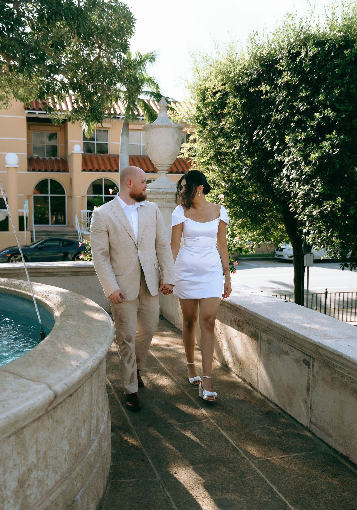 Wedding couple walking along stone wall with Mediterranean architecture in background.