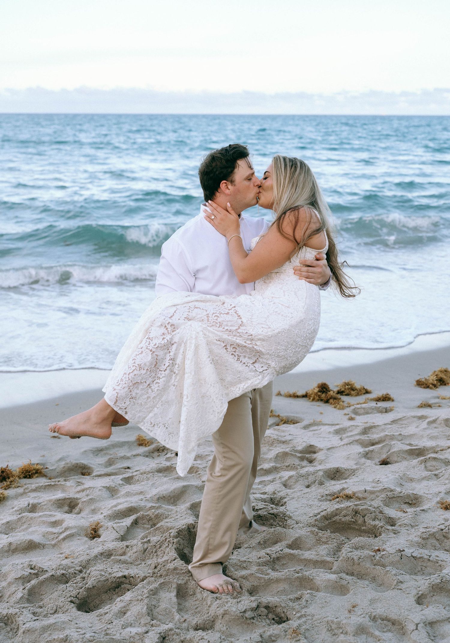 Man lifting woman in a romantic beach kiss with ocean waves behind them.
