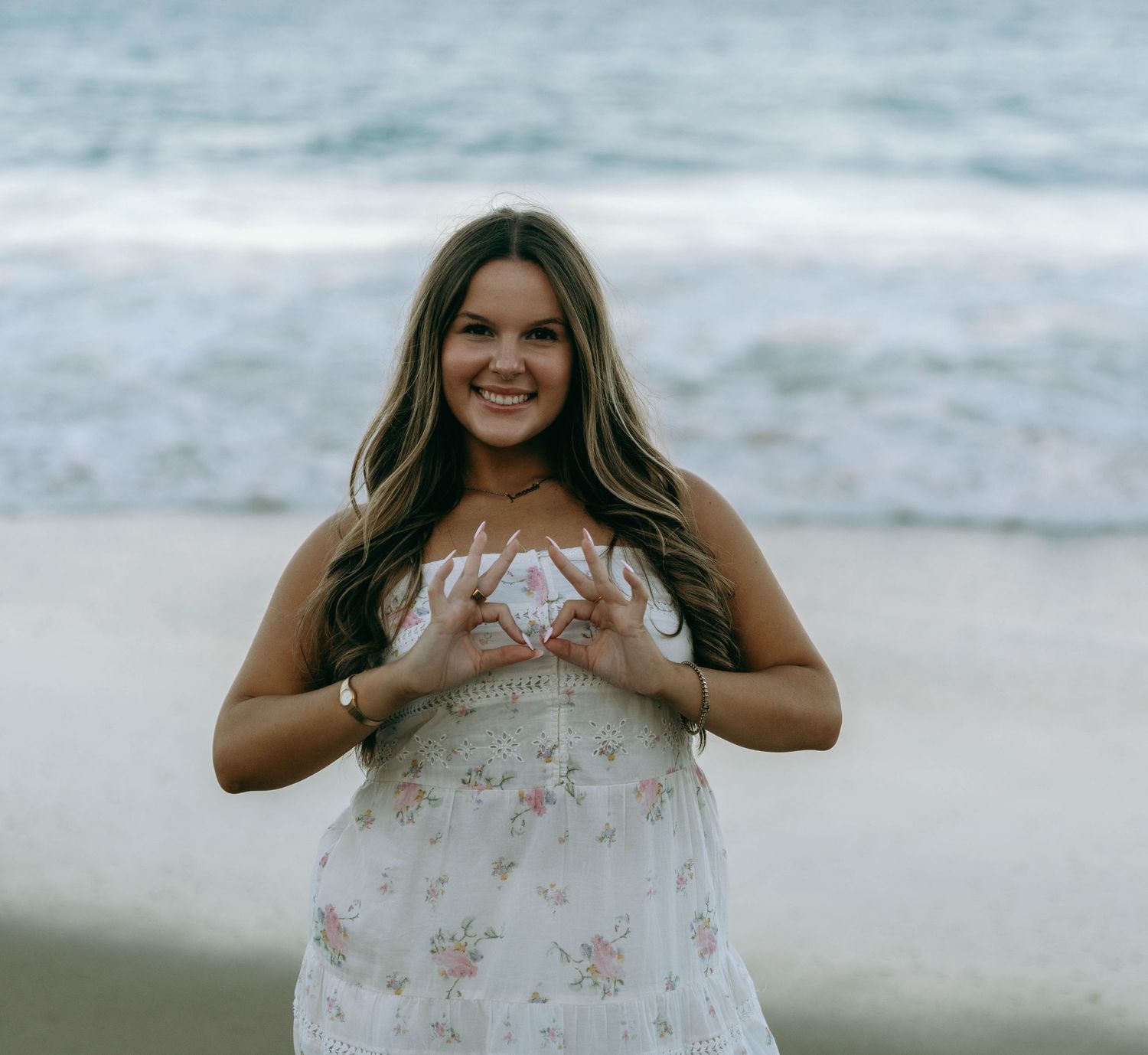 Smiling young woman in floral sundress makes heart shape with hands on sandy beach. Worth Avenue, Palm Beach, FL
