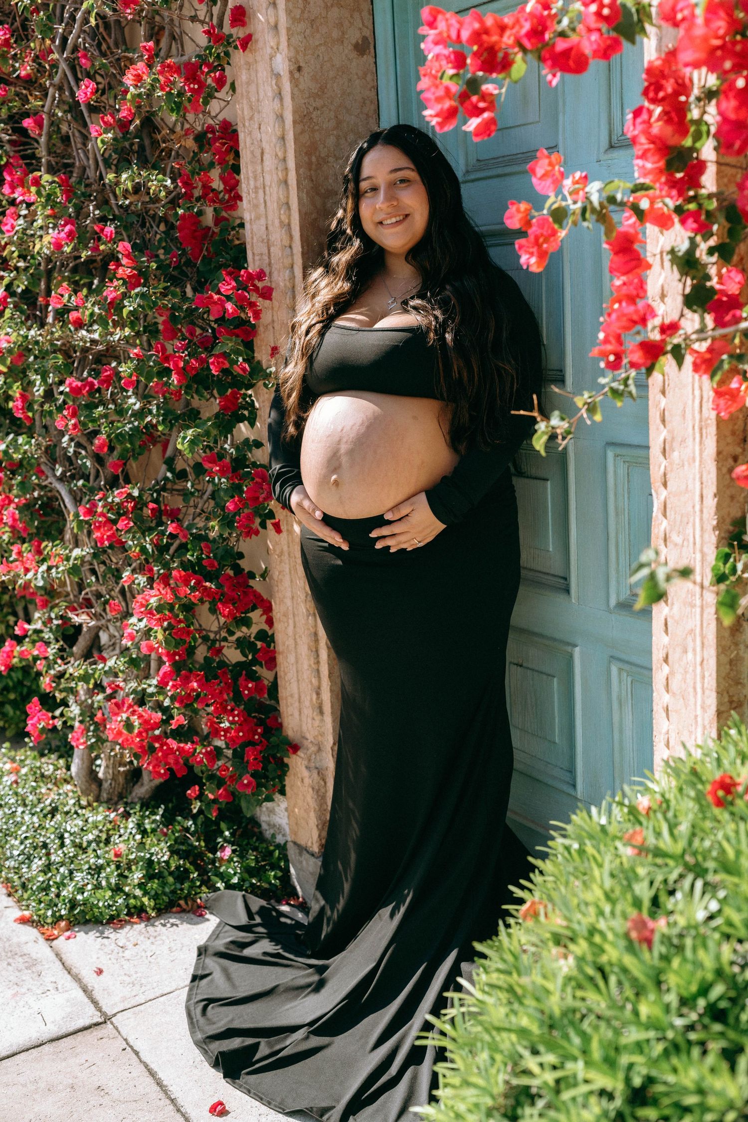 Pregnant woman in black crop top and skirt poses by teal door with red bougainvillea flowers. Worth Avenue, Palm Beach, FL