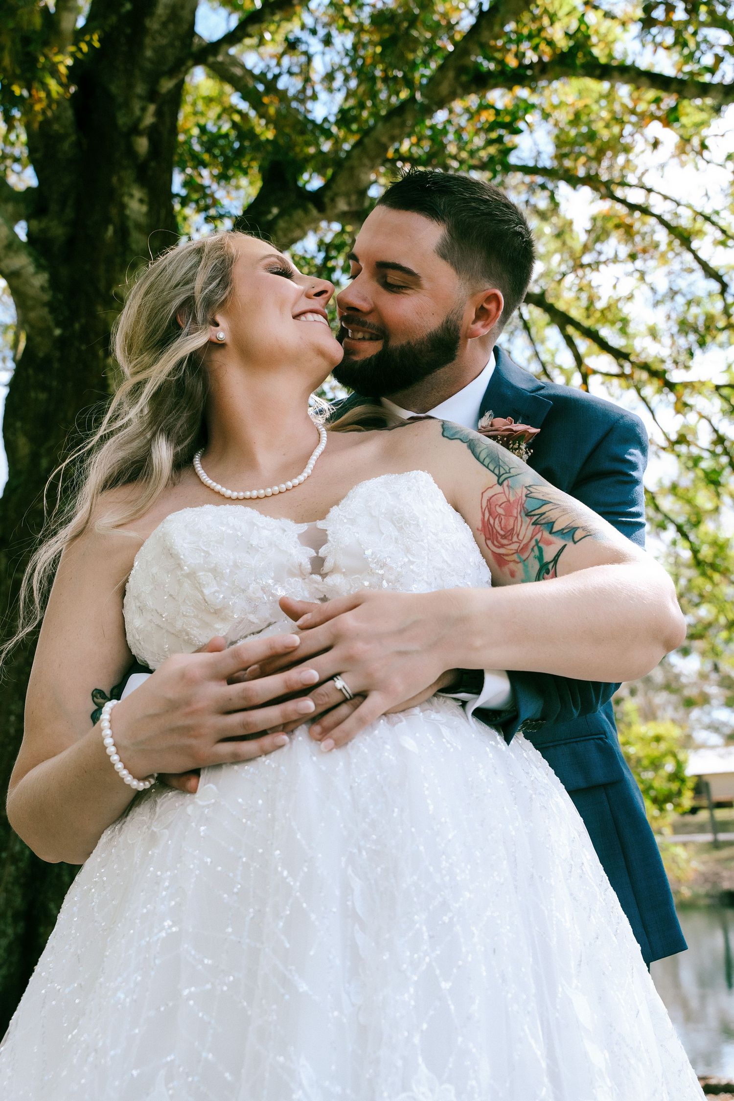 Bearded groom in navy suit embracing bride in white strapless gown outdoors under a tree.