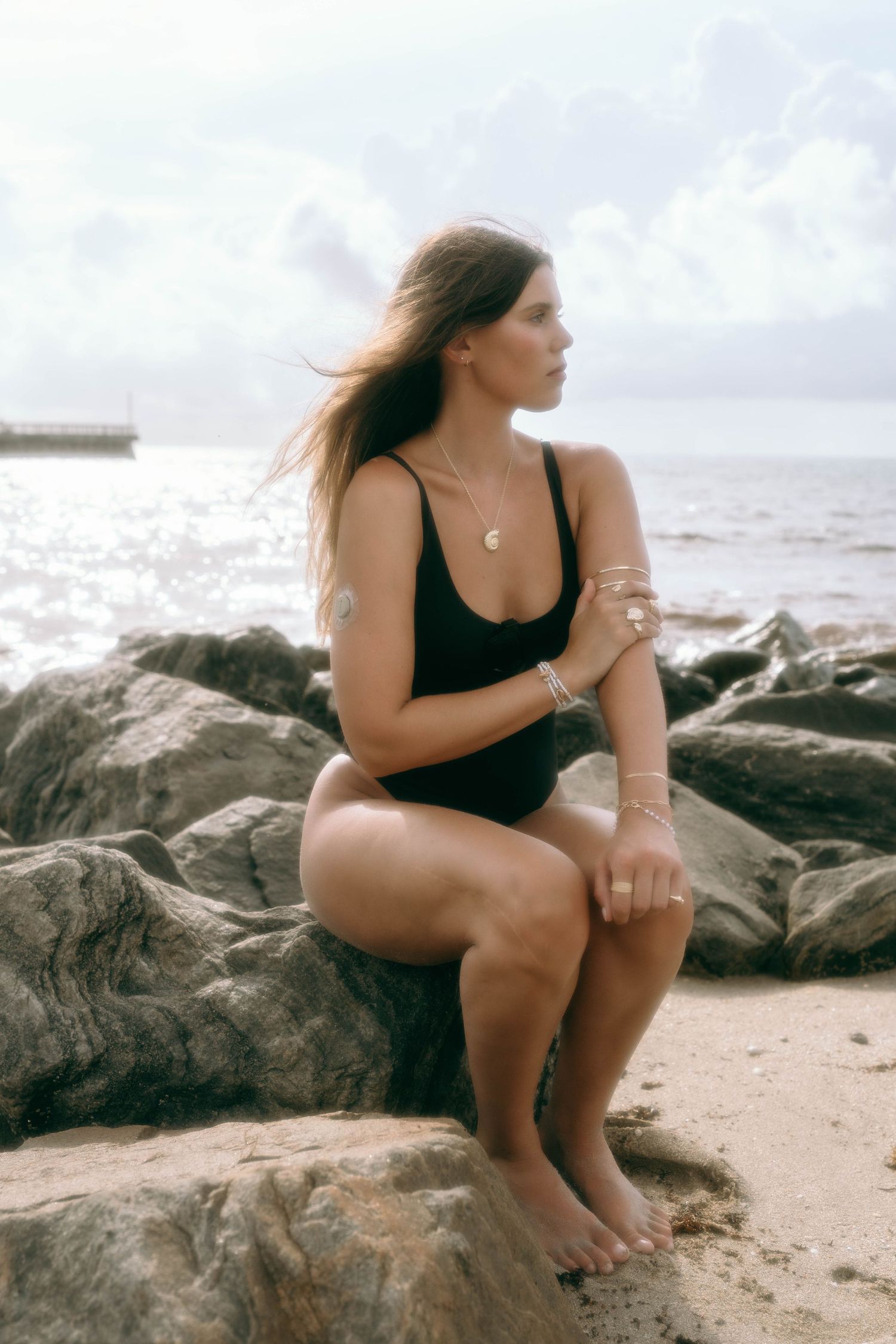 Woman in black one-piece swimsuit sitting on beach rocks with ocean backdrop and wind-blown hair.