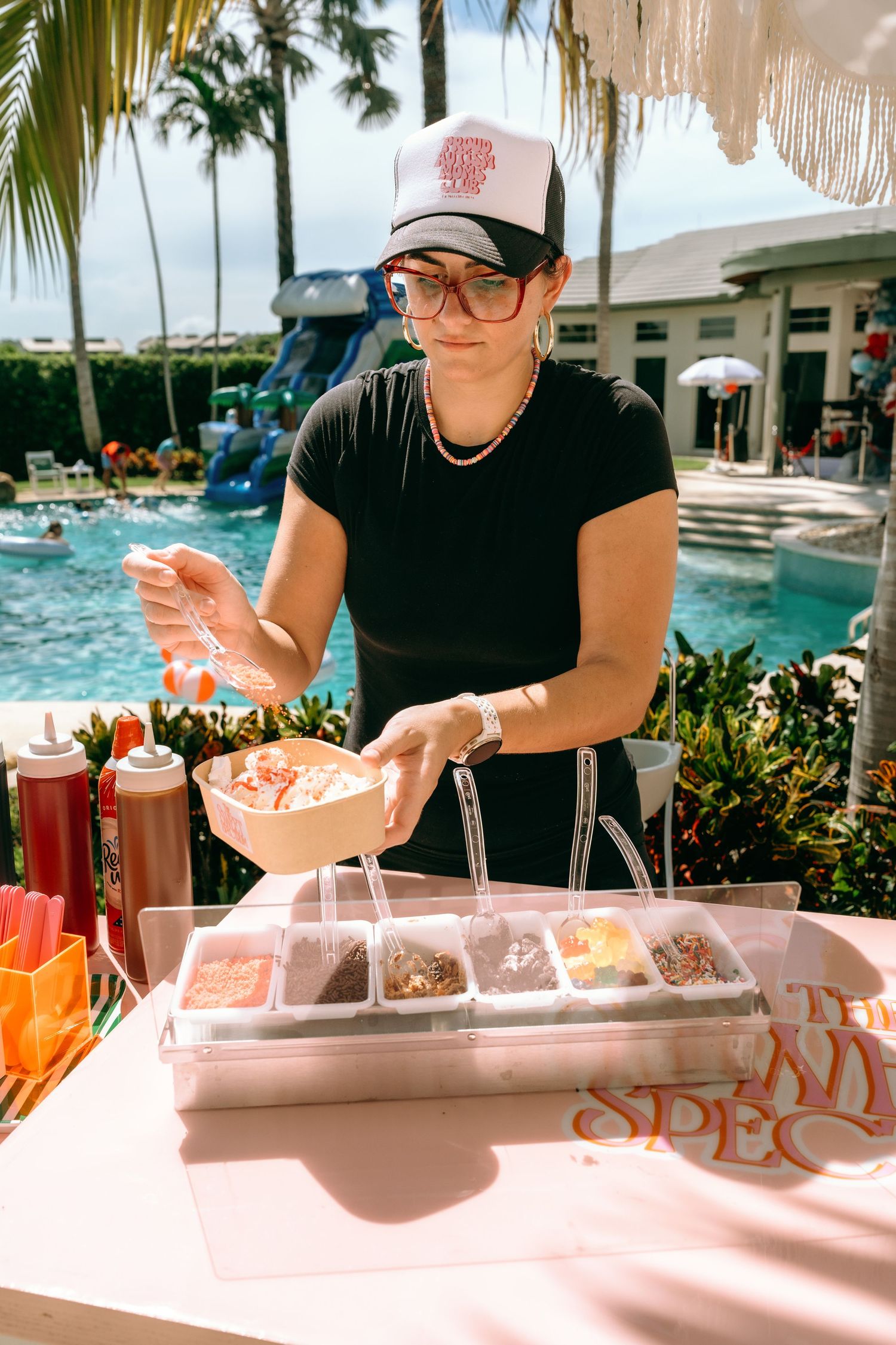 A woman in a black shirt and trucker hat prepares colorful toppings at an outdoor pool party food station.