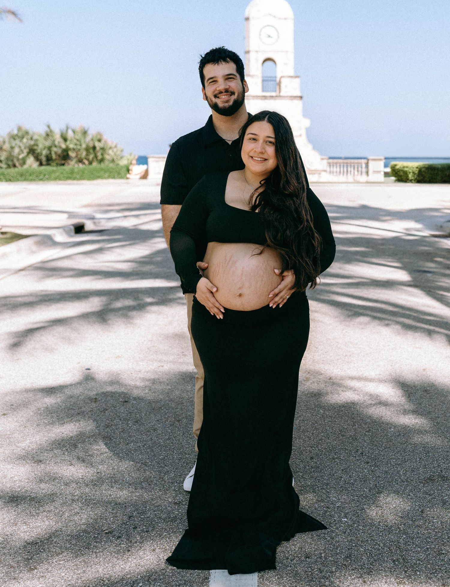 Couple posing for maternity photos outdoors near a clock tower, woman in black dress showing baby bump. Worth Avenue, Palm Beach, FL
