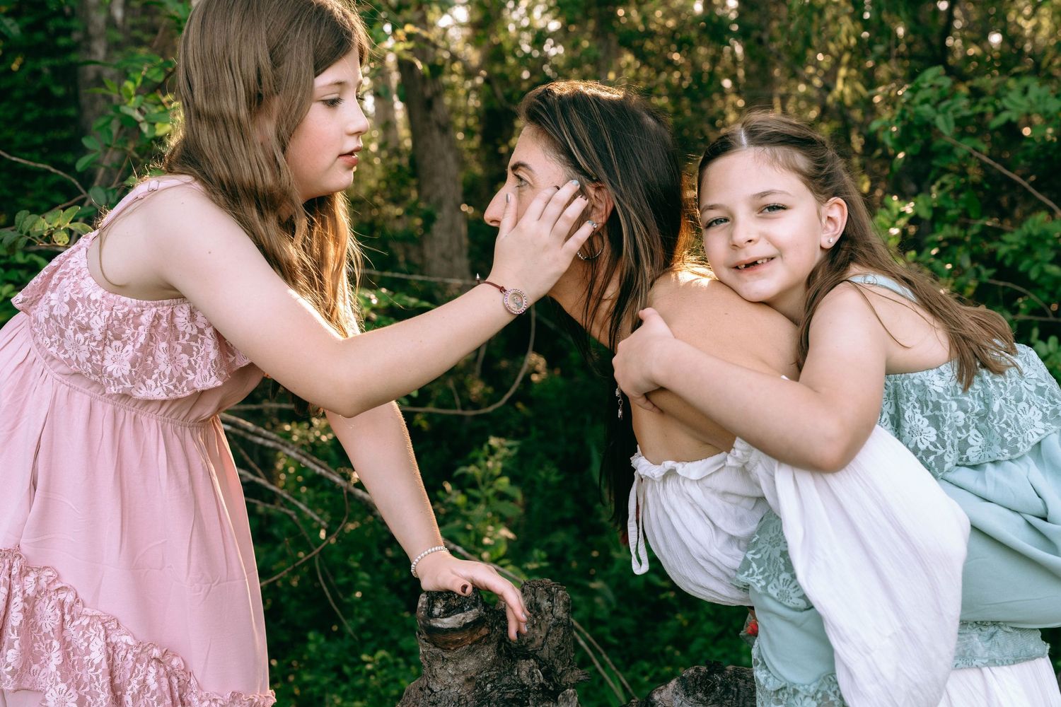 Three young girls playing outdoors among green trees, one whispering while another smiles and hugs her friend. Colt Farms