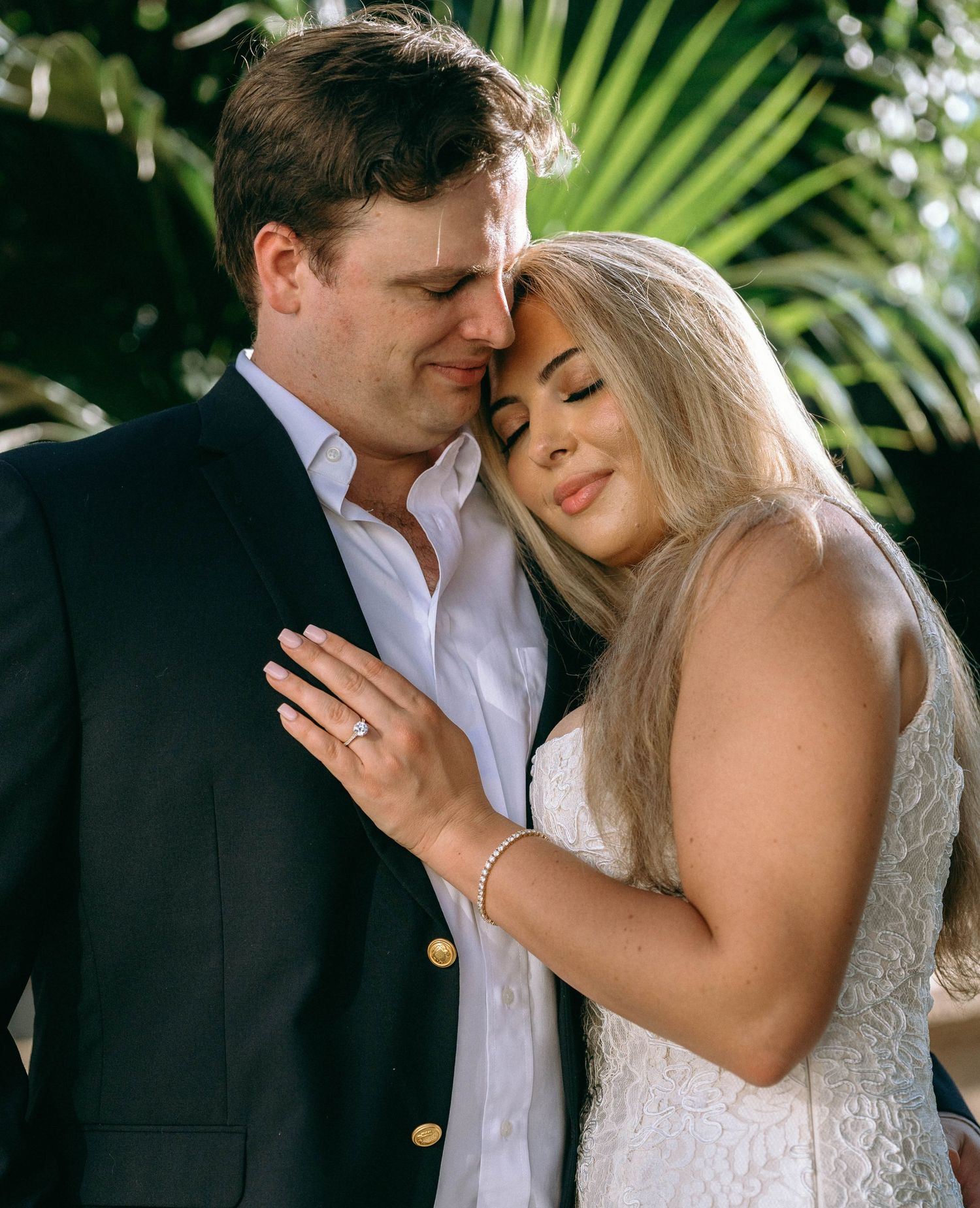 Couple in formal attire sharing intimate moment, bride in white lace dress resting head on grooms chest.