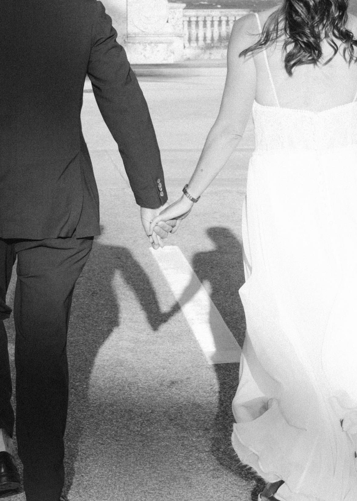Black and white photo of bride and groom holding hands, their shadows forming a heart shape on the ground.