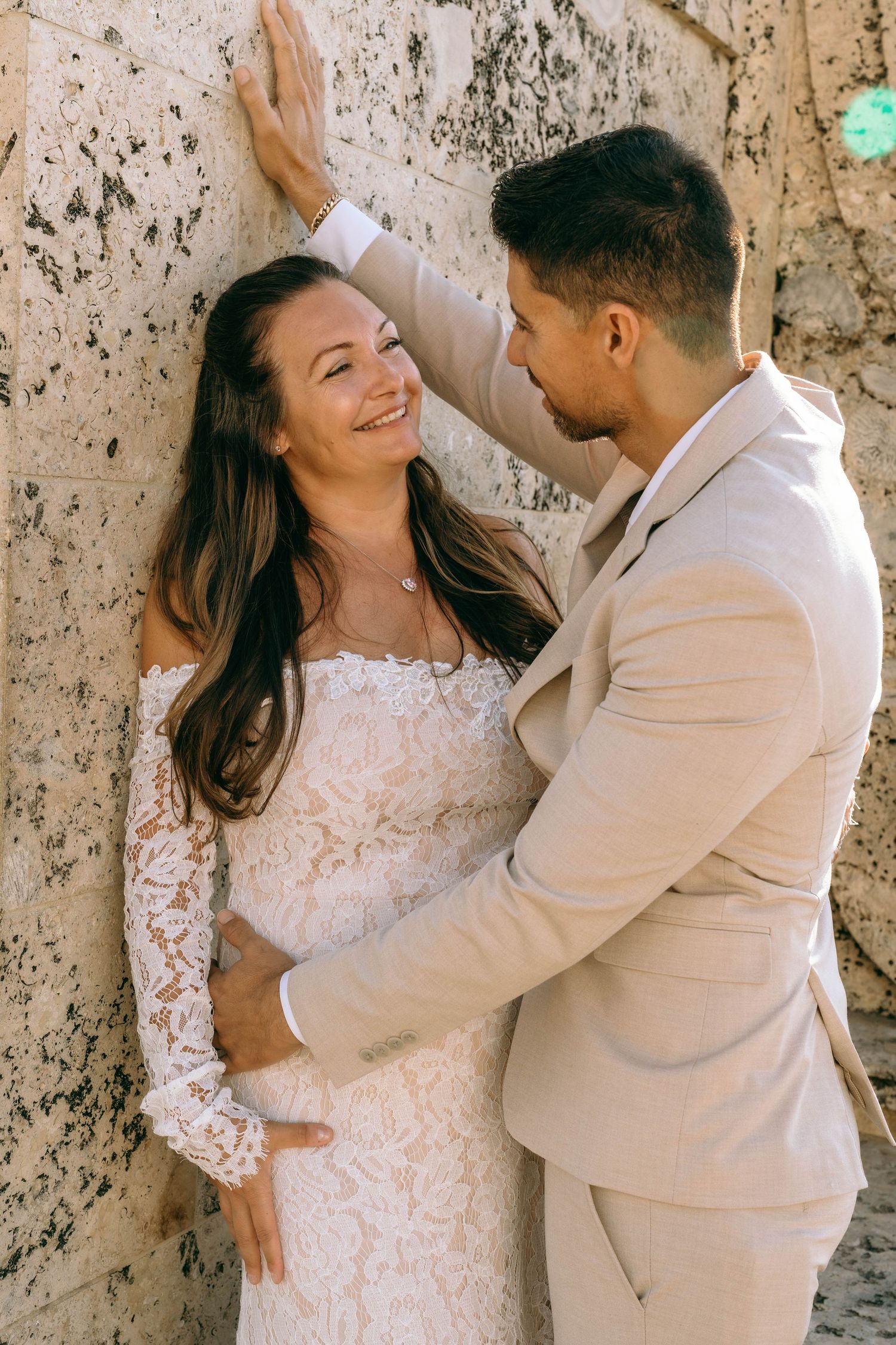 Smiling bride in lace gown leans against stone wall as groom in tan suit gazes at her.