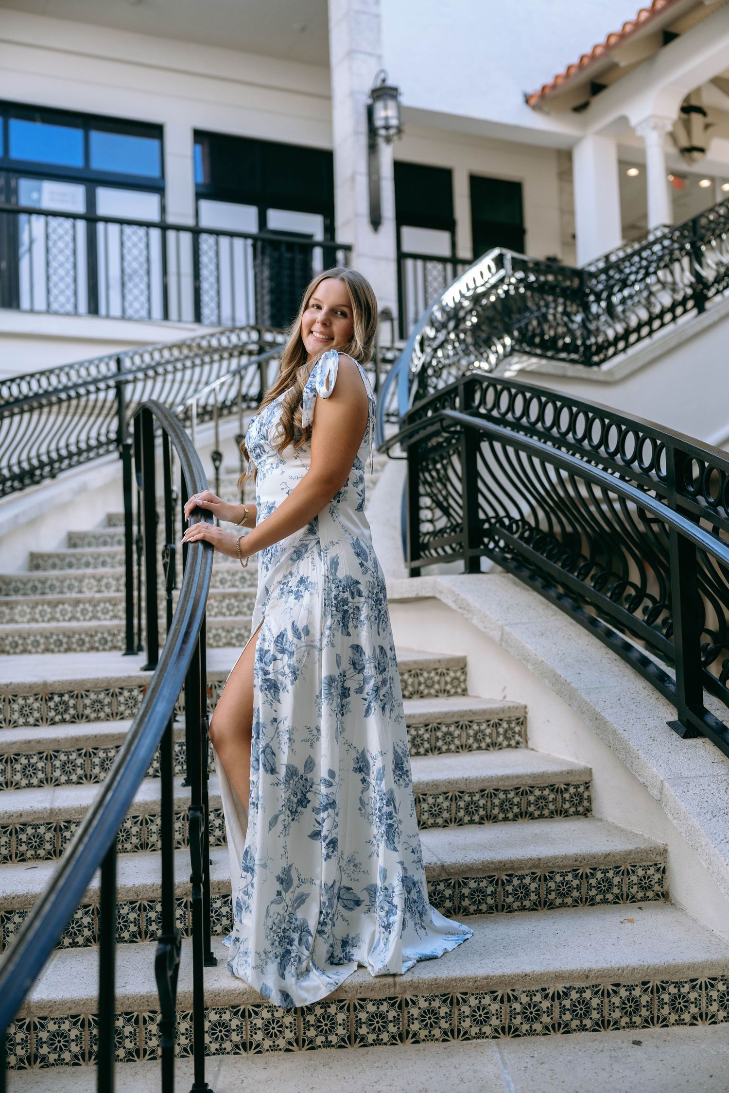 Woman in blue floral maxi dress posing on elegant outdoor staircase with iron railing.