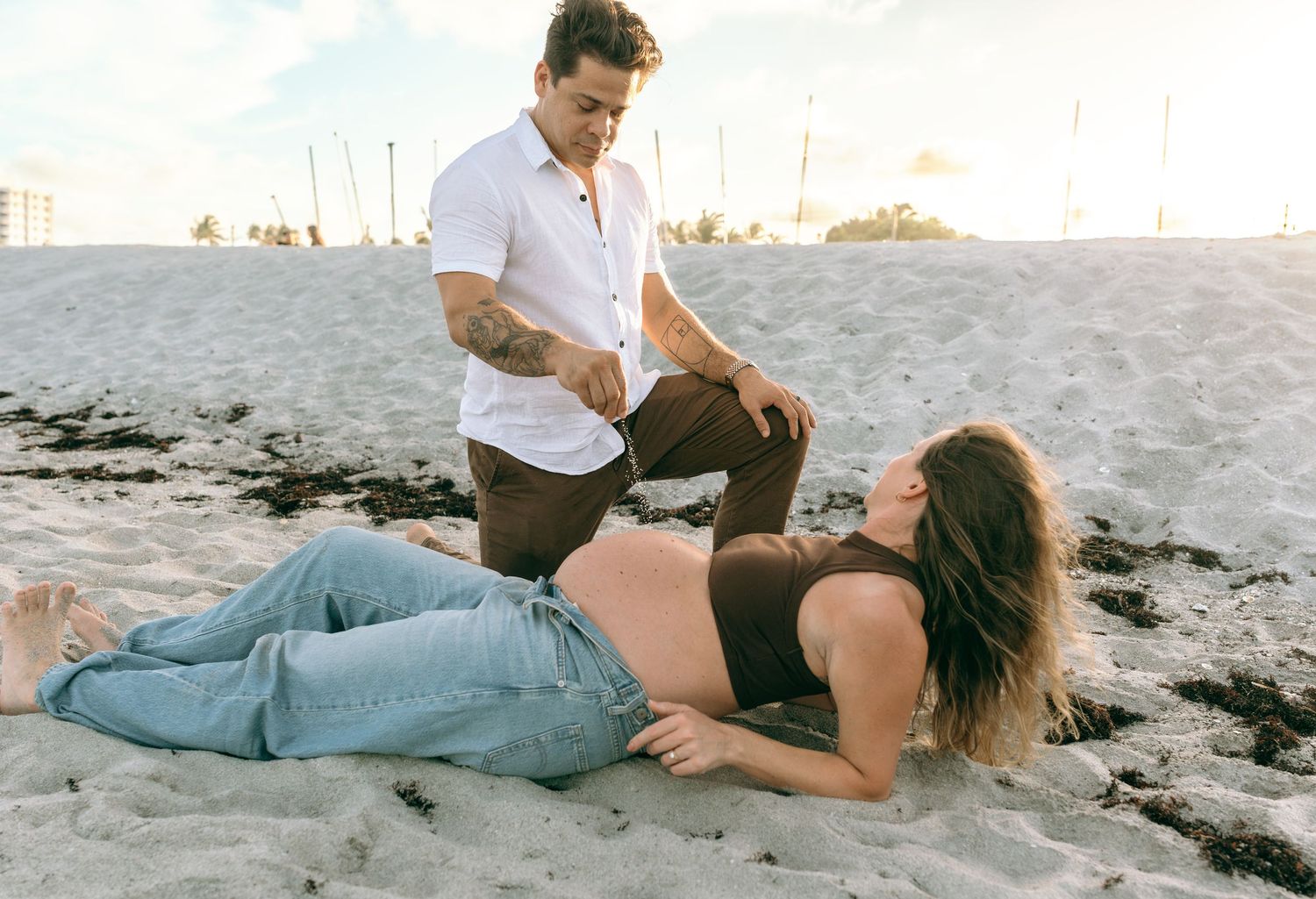 Pregnant woman lying on beach sand while partner stands beside her during a maternity photoshoot at sunset.