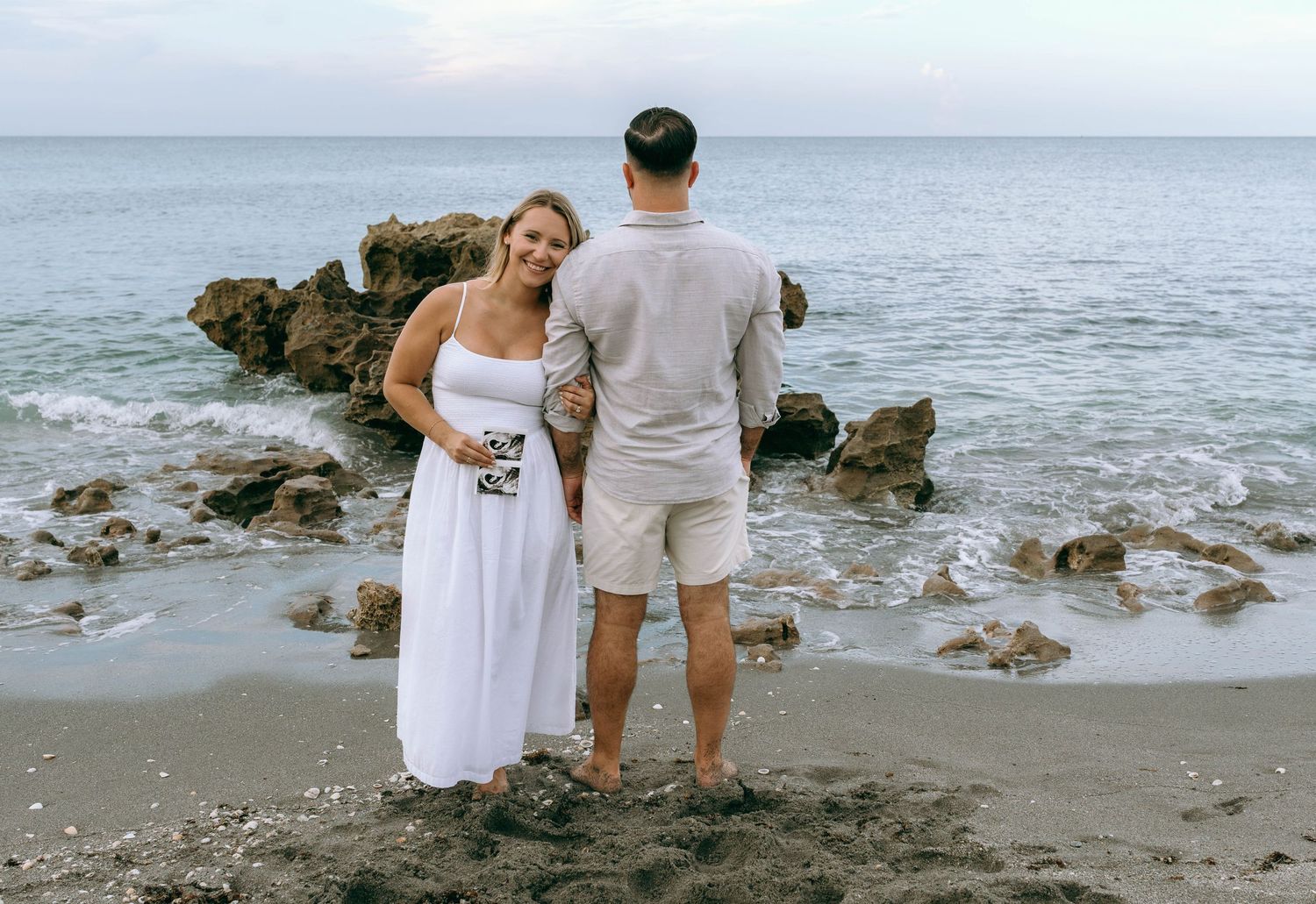 Couple in white outfits standing on a rocky beach by the ocean, woman in white dress smiling at camera.