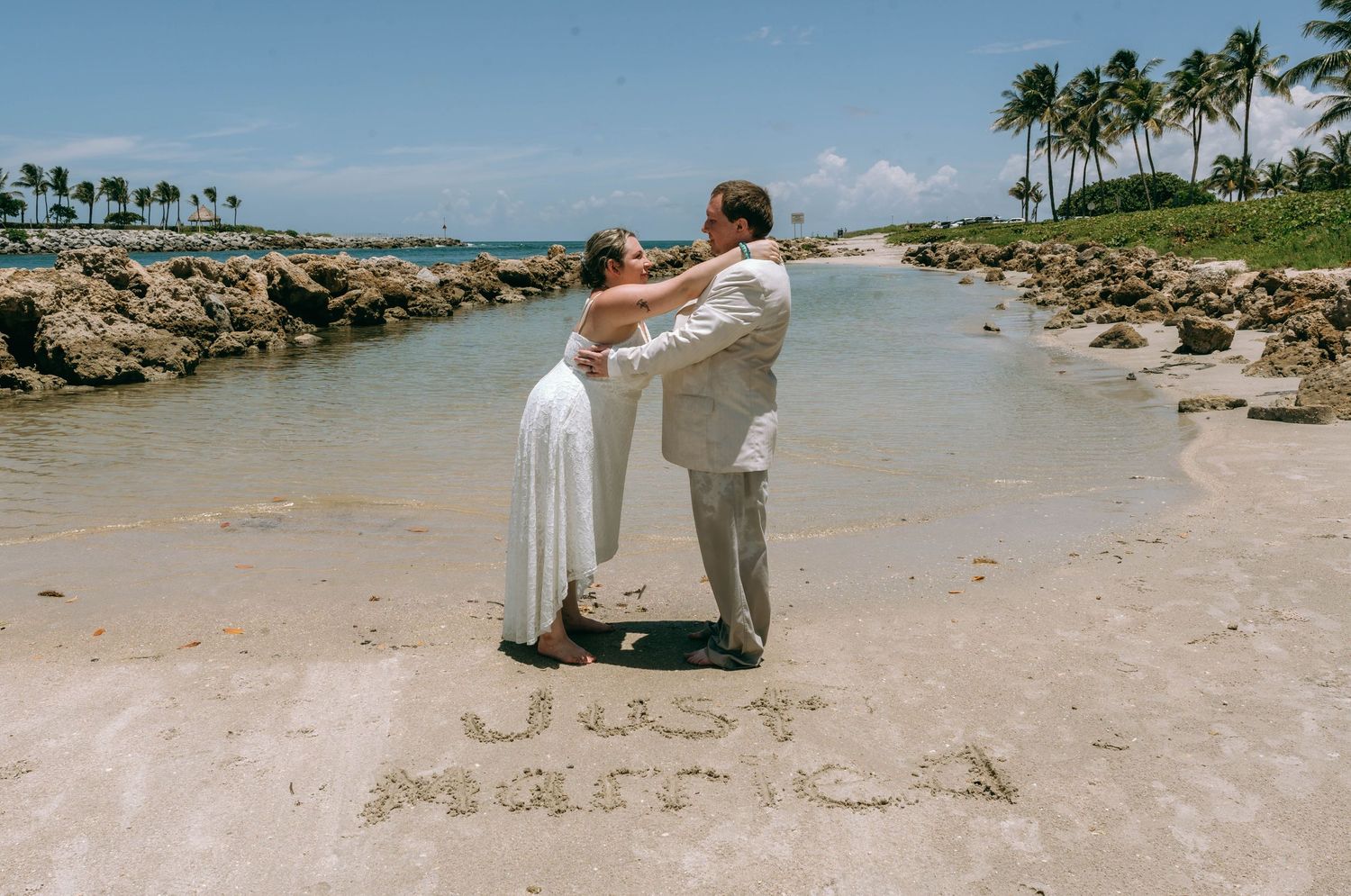 Newlyweds embrace on a tropical beach with Just Married written in sand, surrounded by palm trees and clear skies.