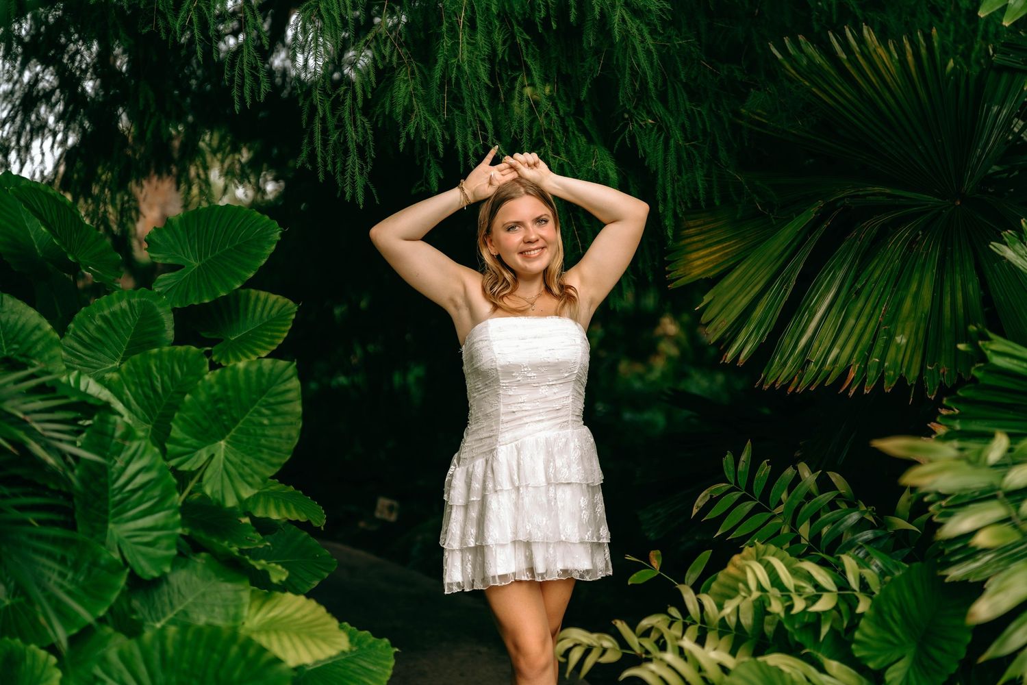 Woman in white mini dress smiling among lush tropical green plants and palm leaves in a garden setting.