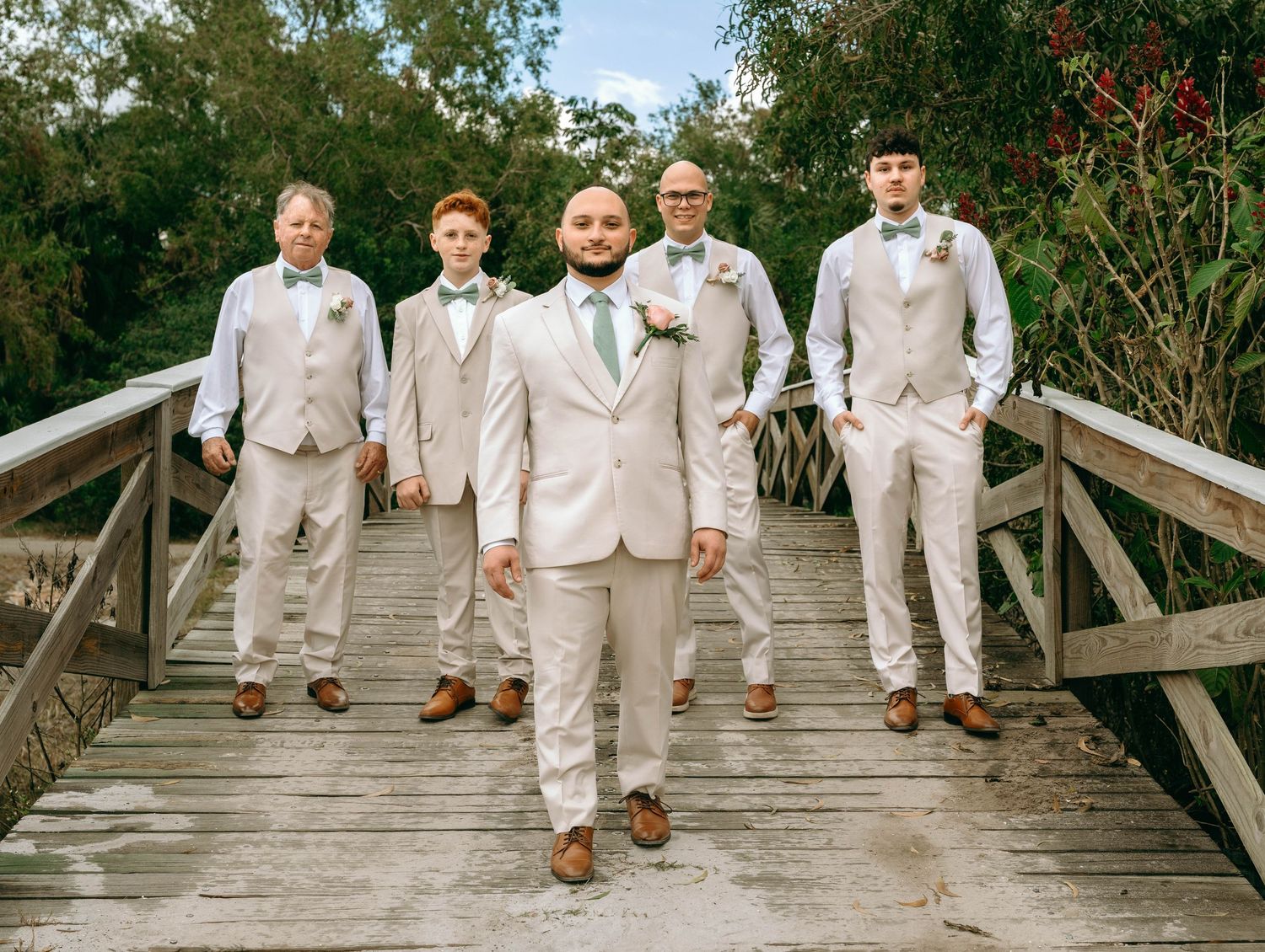 Groomsmen in beige suits and brown shoes sit casually on wooden boardwalk steps during outdoor wedding photoshoot.