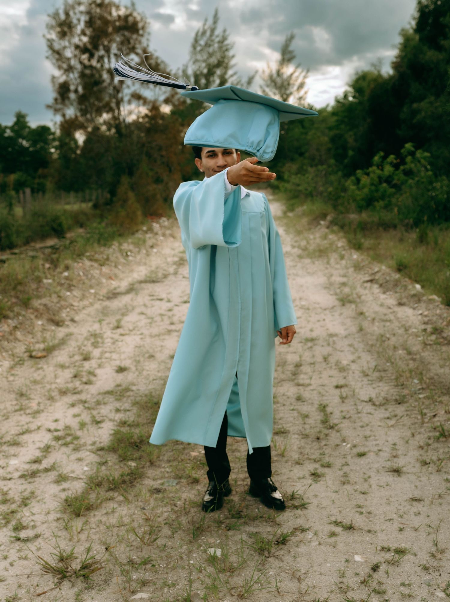 Young child in light blue graduation cap and gown standing on a dirt path, reaching toward camera. Senior Photos at Colt Farms in Boynton