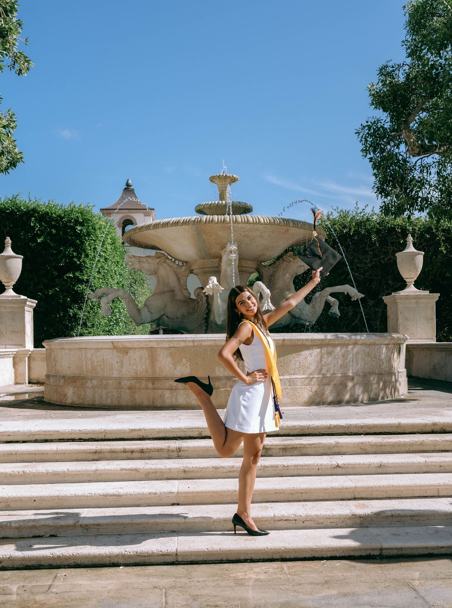 Joyful graduate in white dress with gold sash posing by ornate stone fountain outdoors on Worth Avenue Palm beach.
