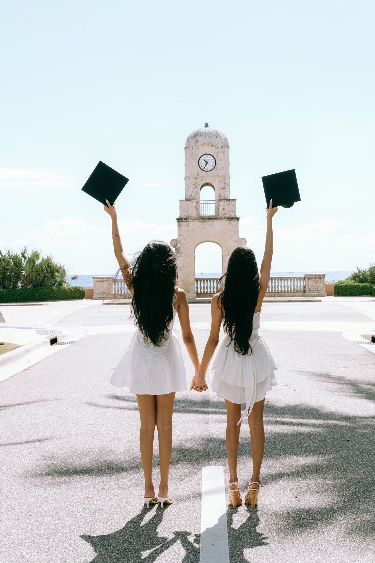 Two women in white dresses holding graduation caps aloft while facing a historic clock tower on Worth Avenue Palm beach.