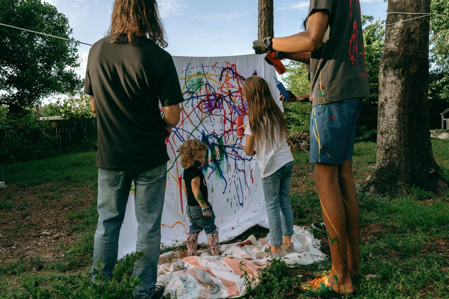Children and adults painting colorful abstract art on a large white canvas outdoors in a green yard.