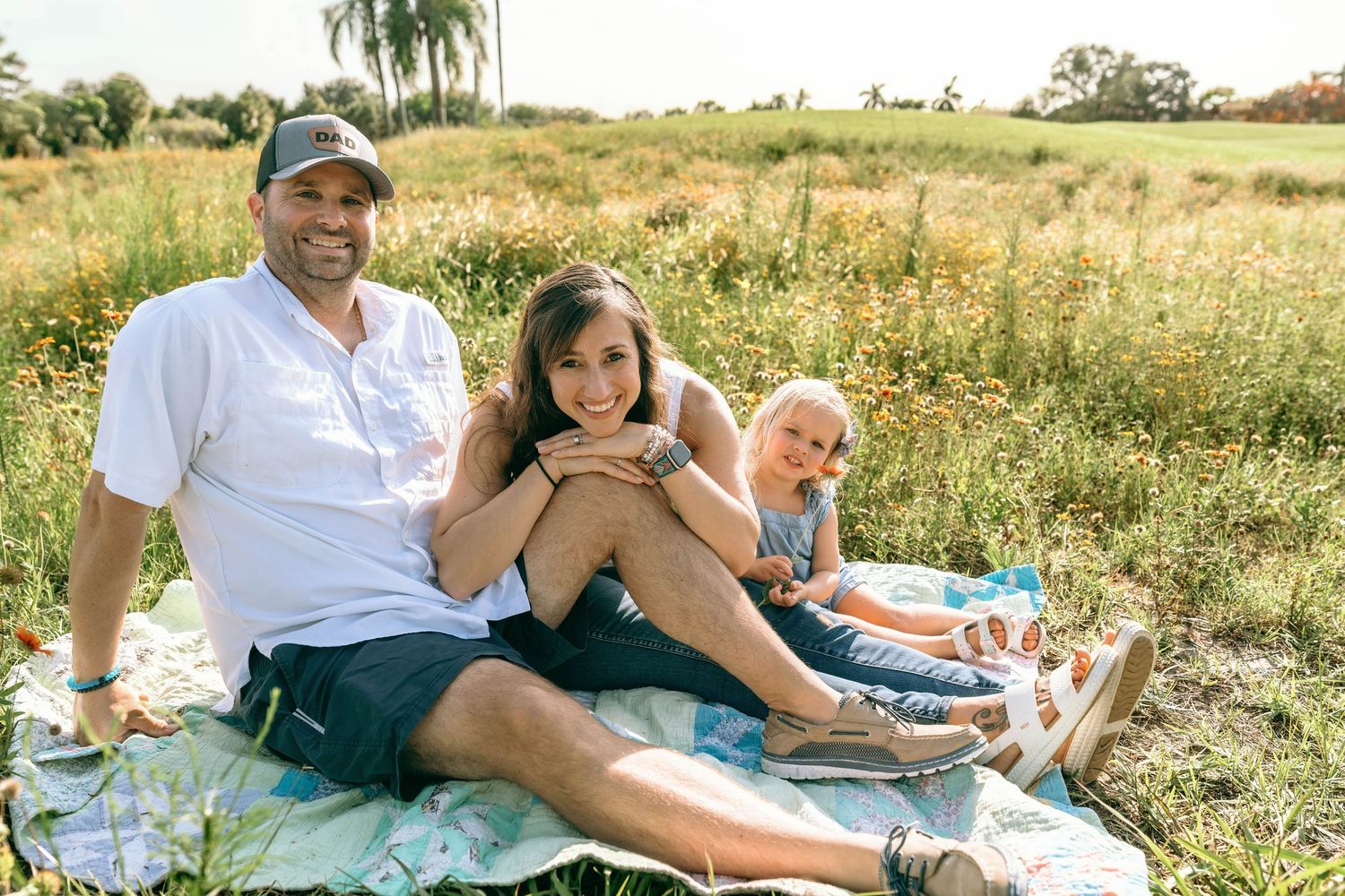 Family of three smiling on a blanket in a sunny meadow, enjoying a relaxed outdoor picnic together.