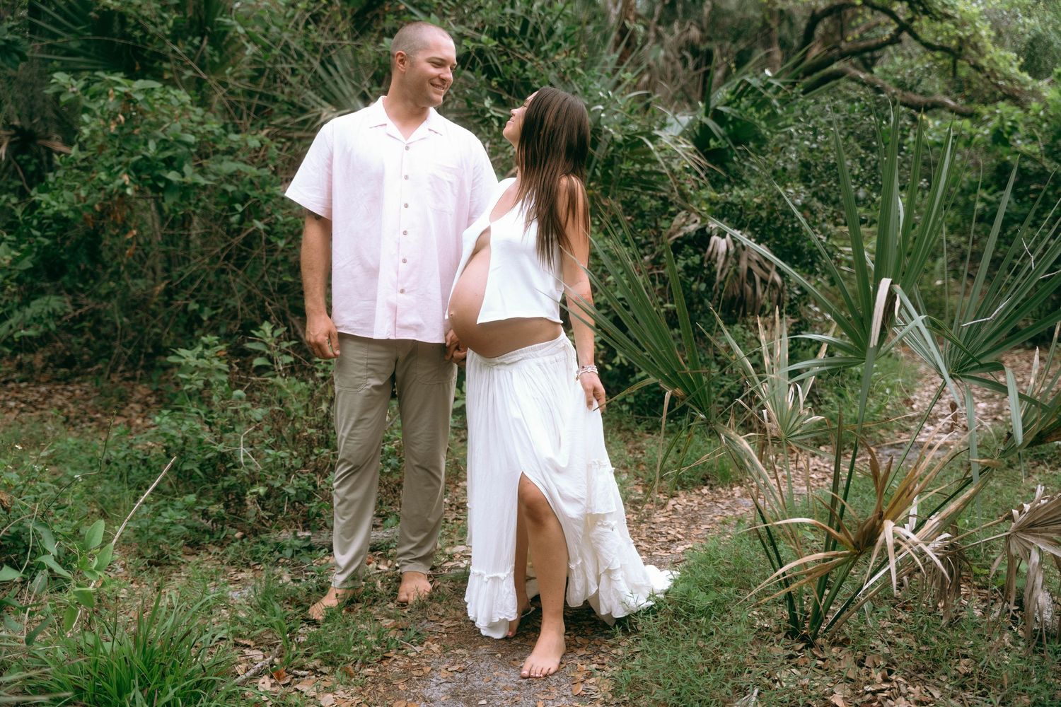 A couple in white clothing walking through a lush green garden path during an outdoor photoshoot.