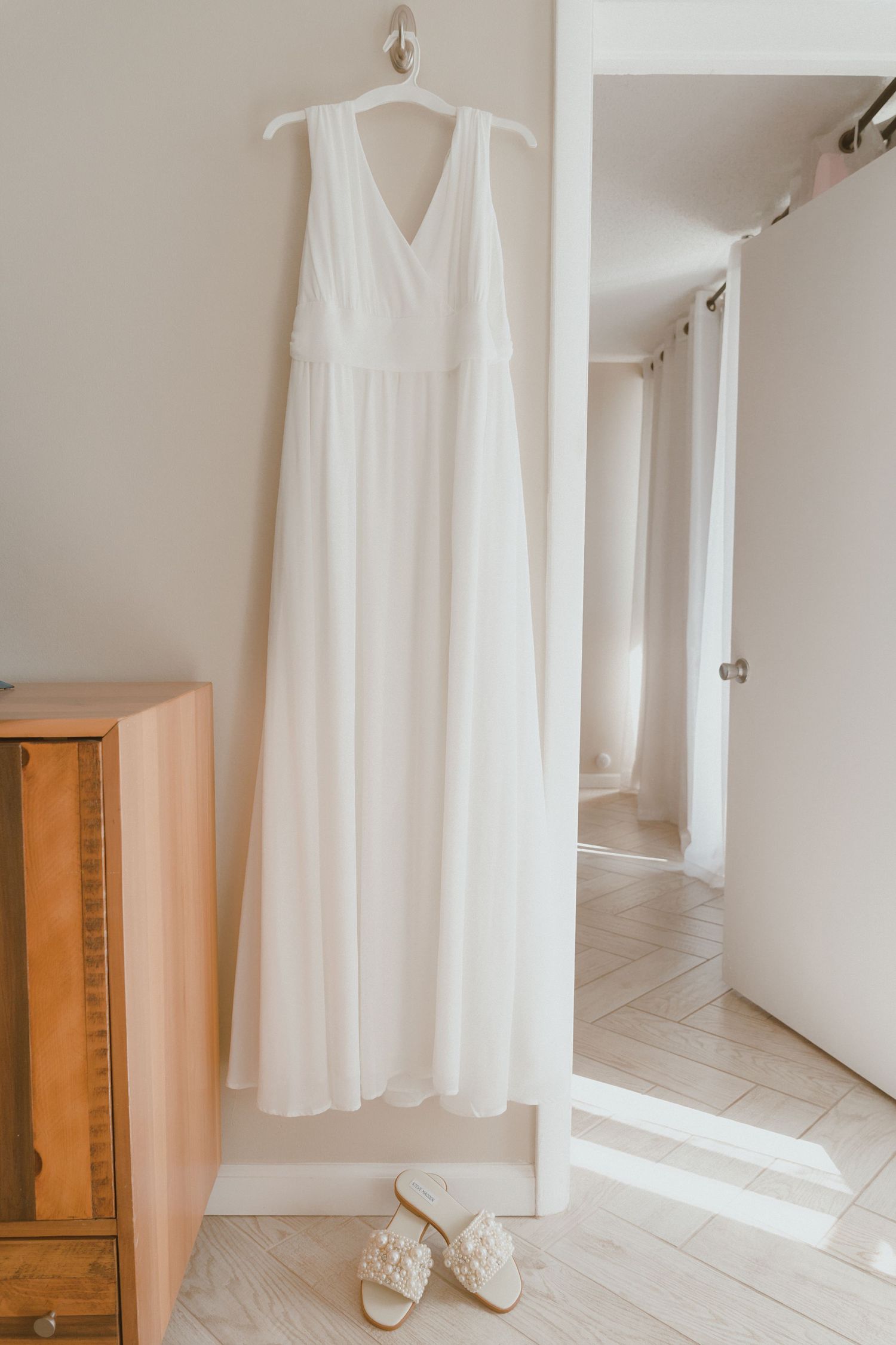 White wedding dress and shoes displayed on hangers against a bright white wall.