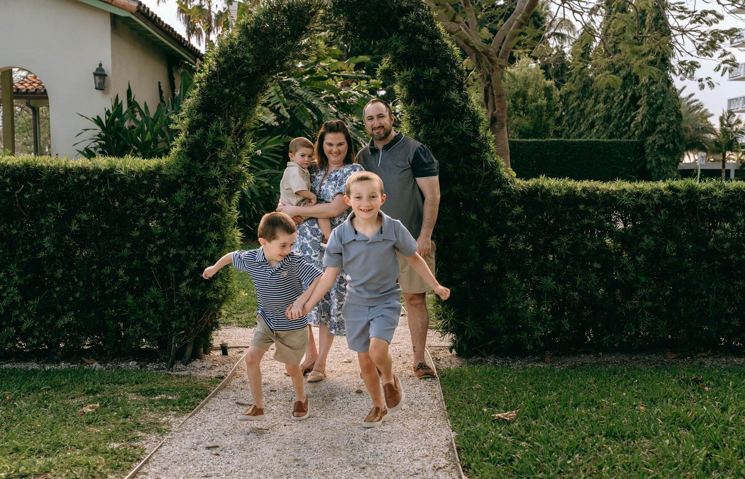 A joyful family runs through a lush garden archway, with two boys in front and parents holding a baby behind.  Family photos at Bradley Park