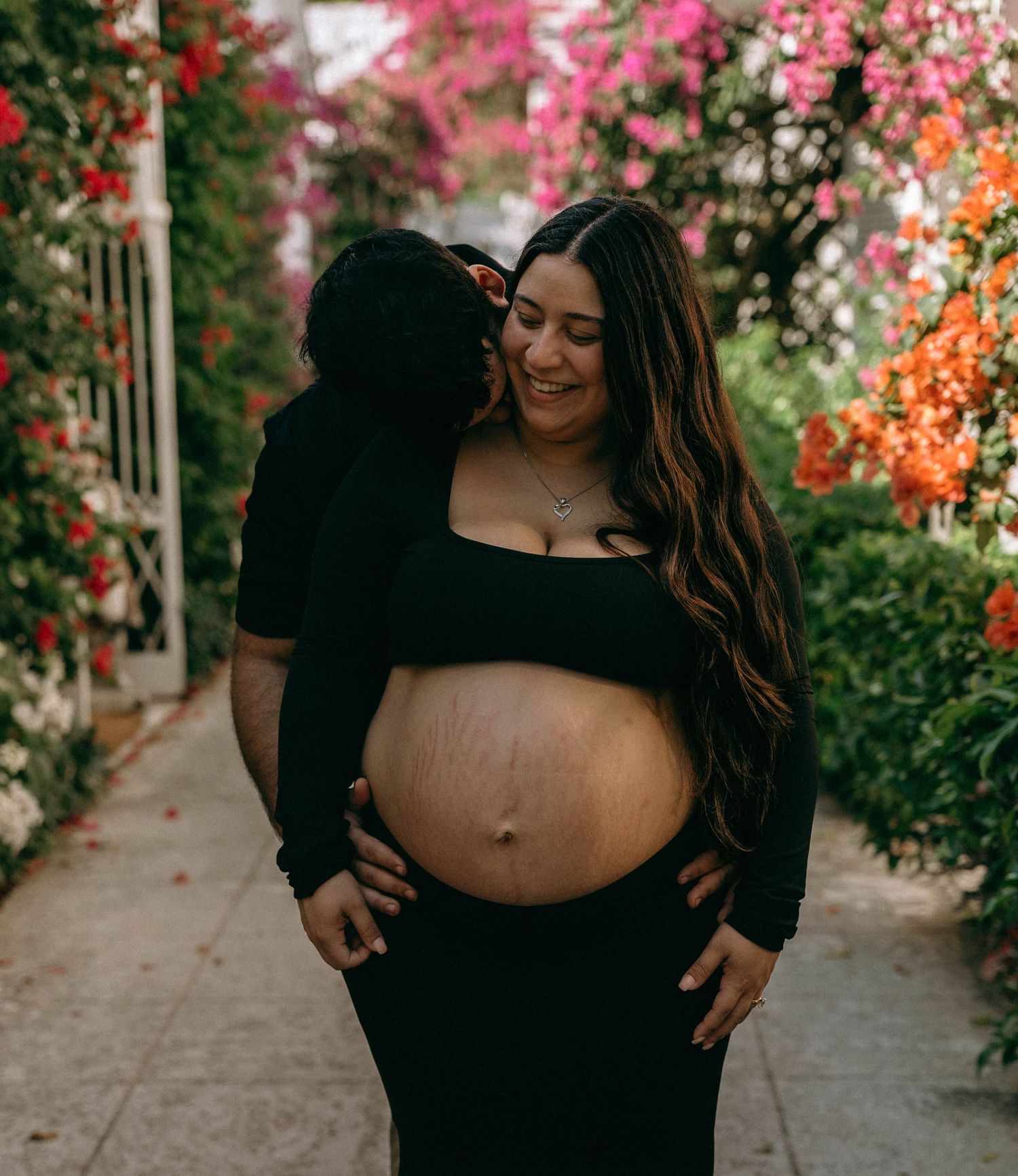Smiling pregnant woman in black crop top embraced by partner among vibrant pink bougainvillea flowers. Worth Avenue, Palm Beach, FL
