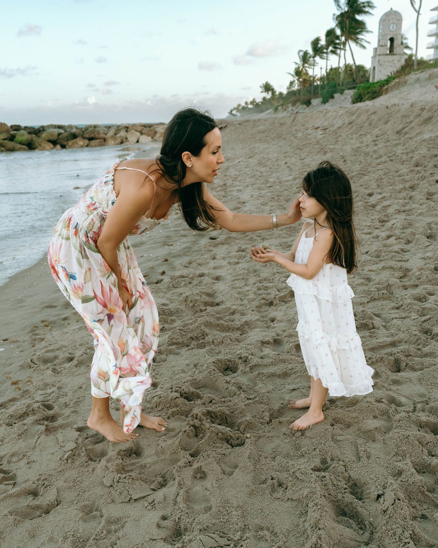 A mother in a floral dress bends to interact with her young daughter in a white dress on a sandy beach at sunset. Worth Avenue, Palm Beach,