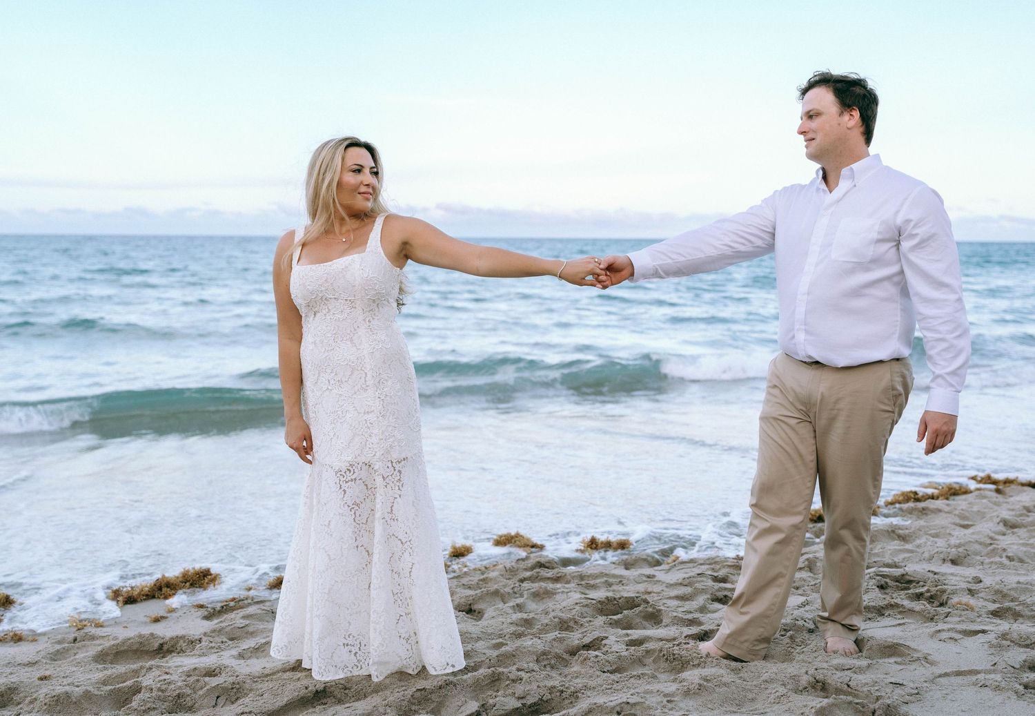 Bride in white lace dress holding grooms hand on sandy beach with ocean waves behind them.