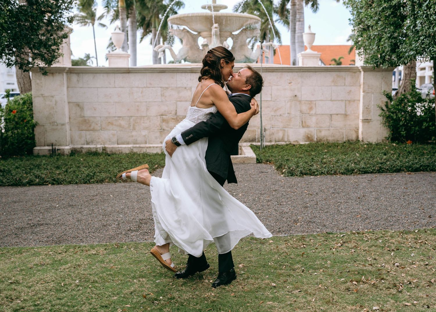 Groom lifting bride in white dress outdoors near fountain, sharing romantic kiss at tropical wedding venue.