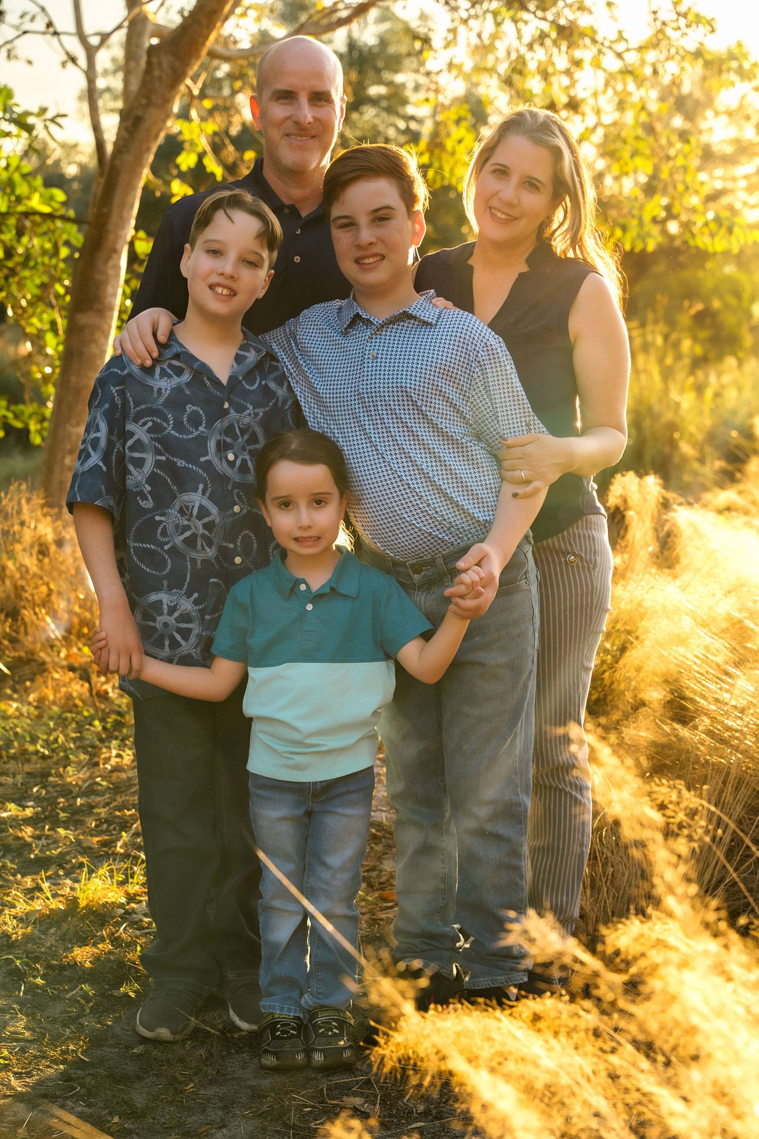 Family of five posing outdoors in golden hour sunlight, with two parents and three boys smiling in a natural setting.