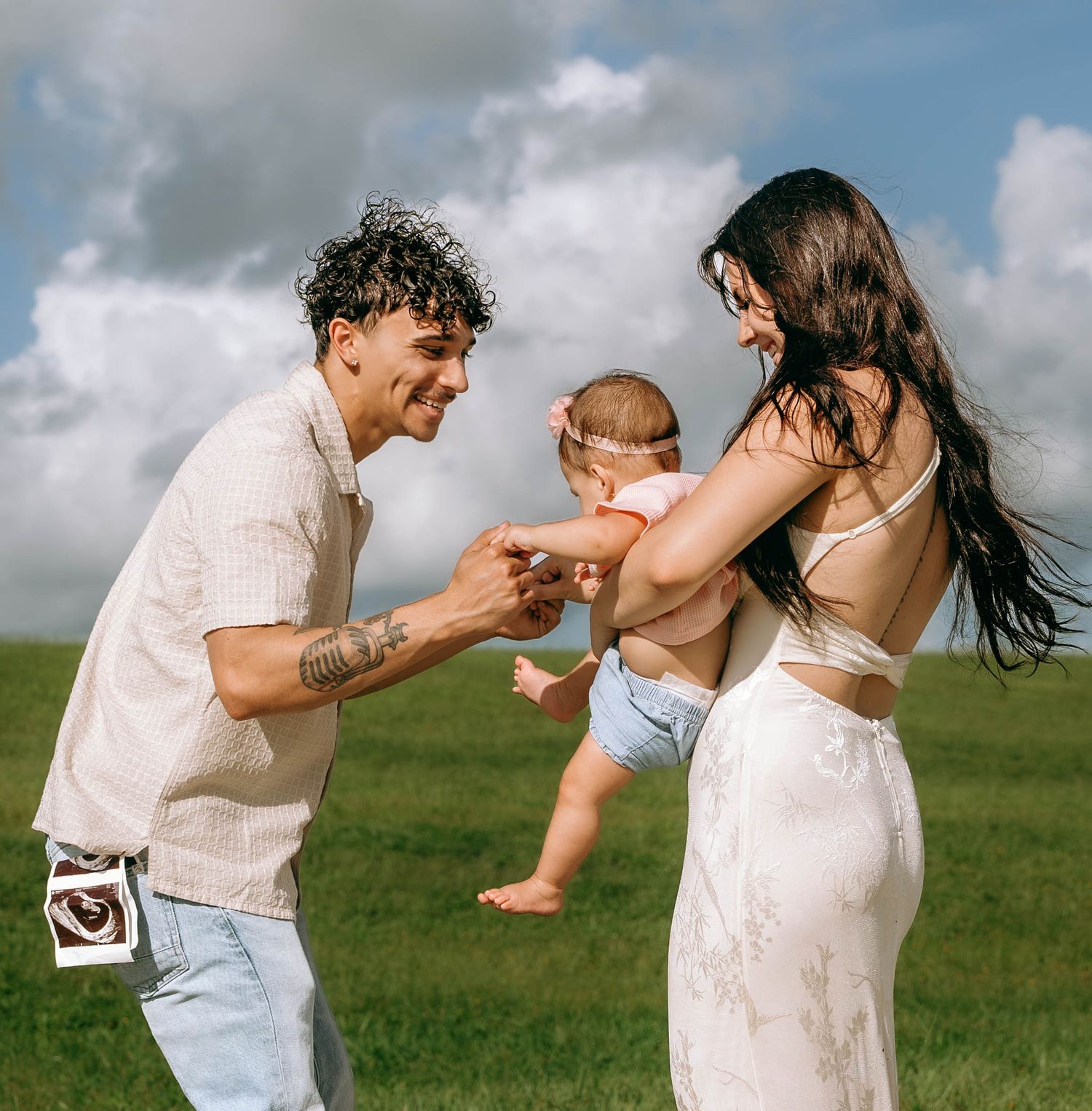 Young couple playing with their baby outdoors on green grass under a cloudy sky on a sunny day.