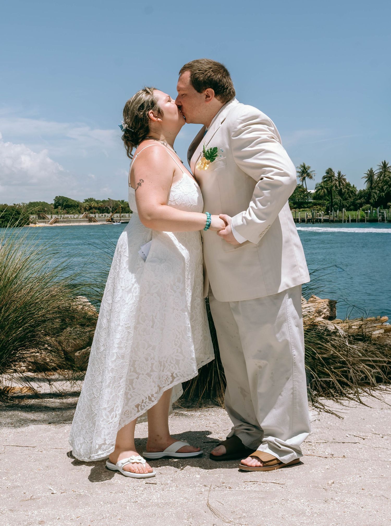 Bride and groom sharing a kiss in wedding attire by a tropical waterfront on a sunny day.