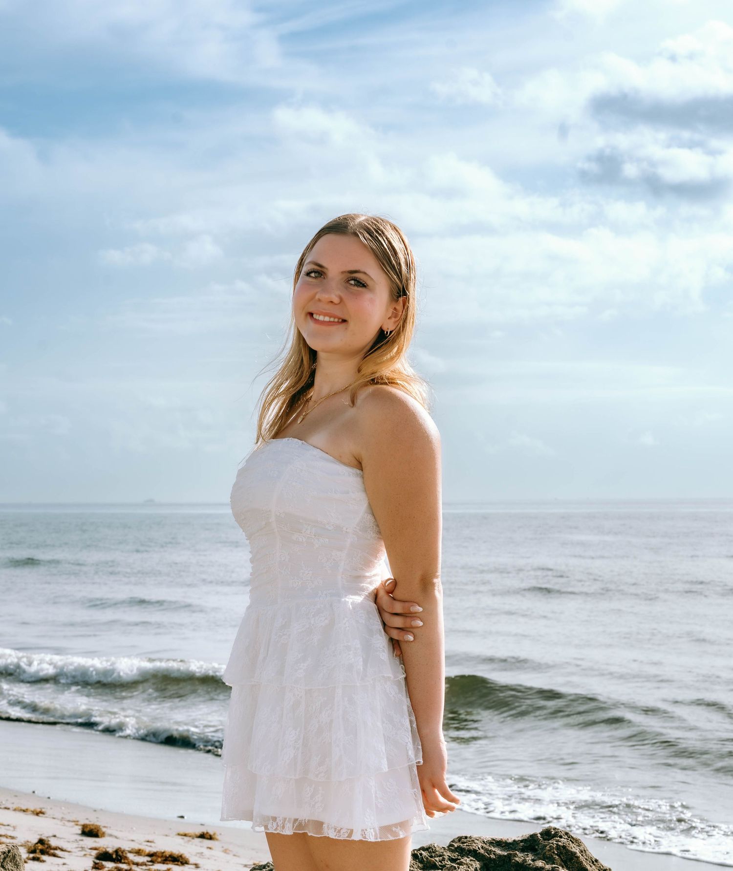 Young woman in white strapless dress smiling on a sunny beach with ocean waves and cloudy sky behind her.