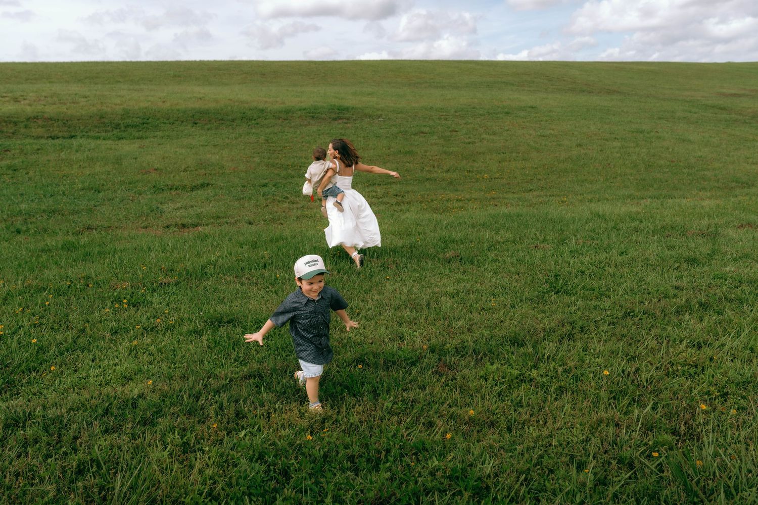 A mother in a white dress and young boy run and play together on a lush green grassy field outdoors.