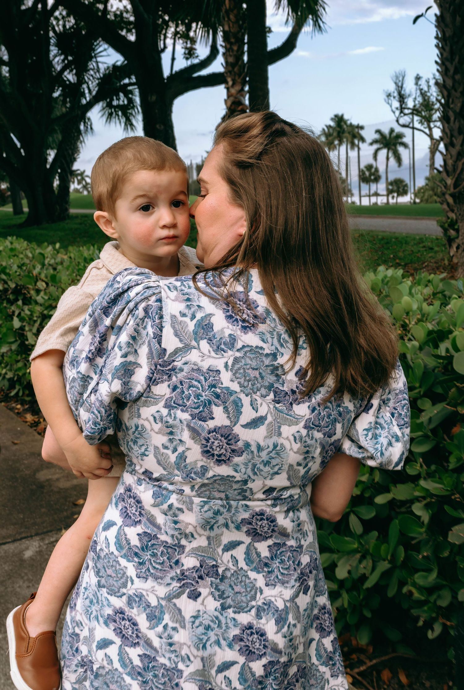A mother kisses her toddler son on the cheek while holding him in a lush outdoor setting. Family photos at Bradley Park Palm Beach, FL