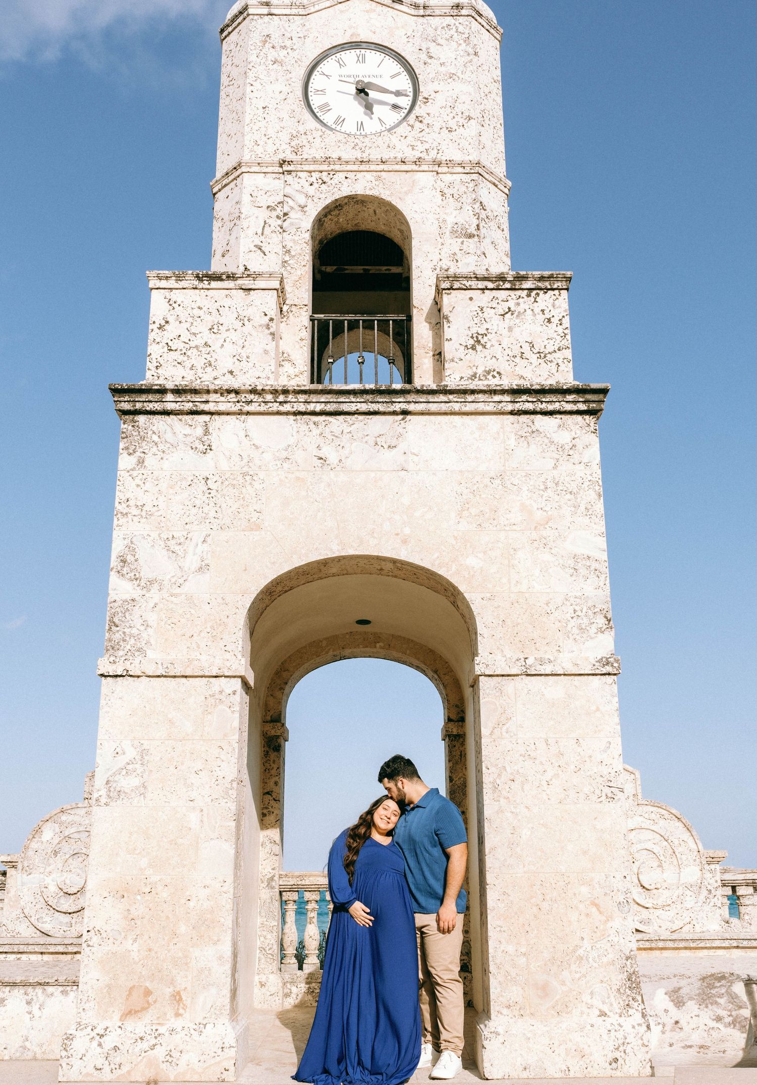 Couple sharing tender moment under historic clock tower arch in blue maternity dress outdoors. Worth Avenue, Palm Beach, FL