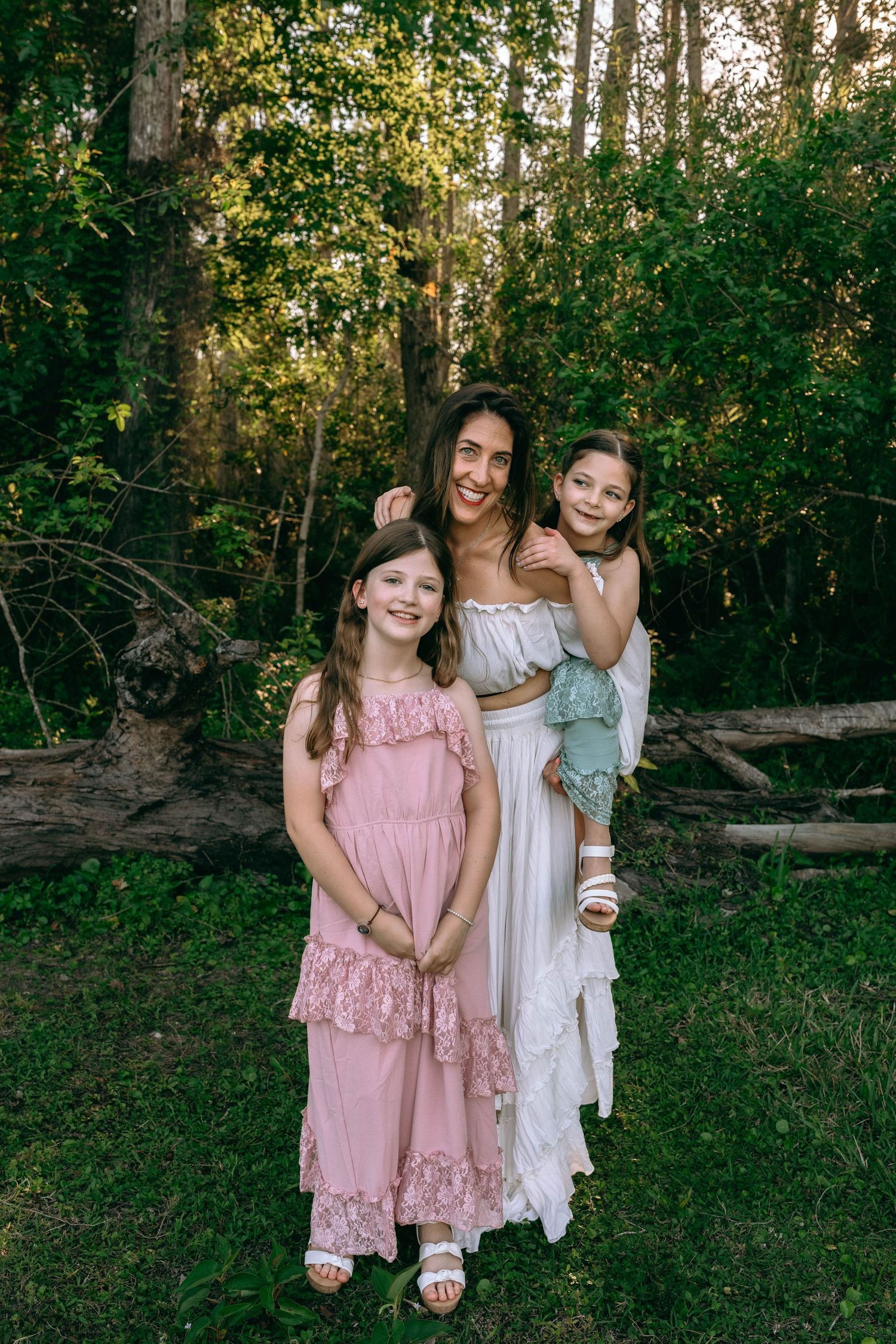 A mother in a white dress holds her daughter while another girl in pink stands beside them outdoors. Colt Farms
