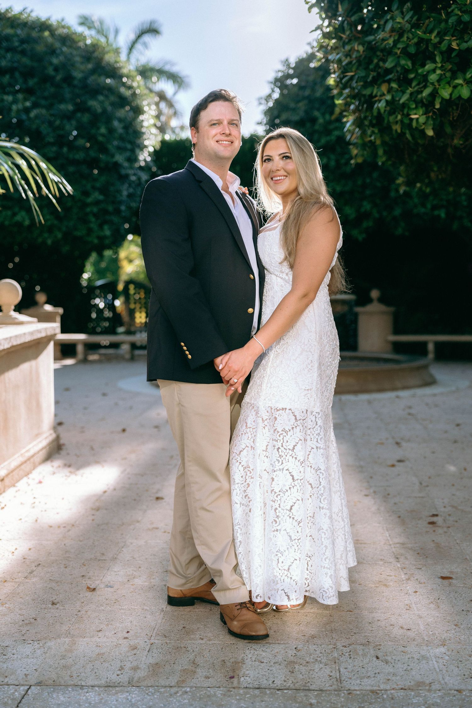 Smiling couple holding hands in a sunlit garden, man in blazer, woman in white lace dress.