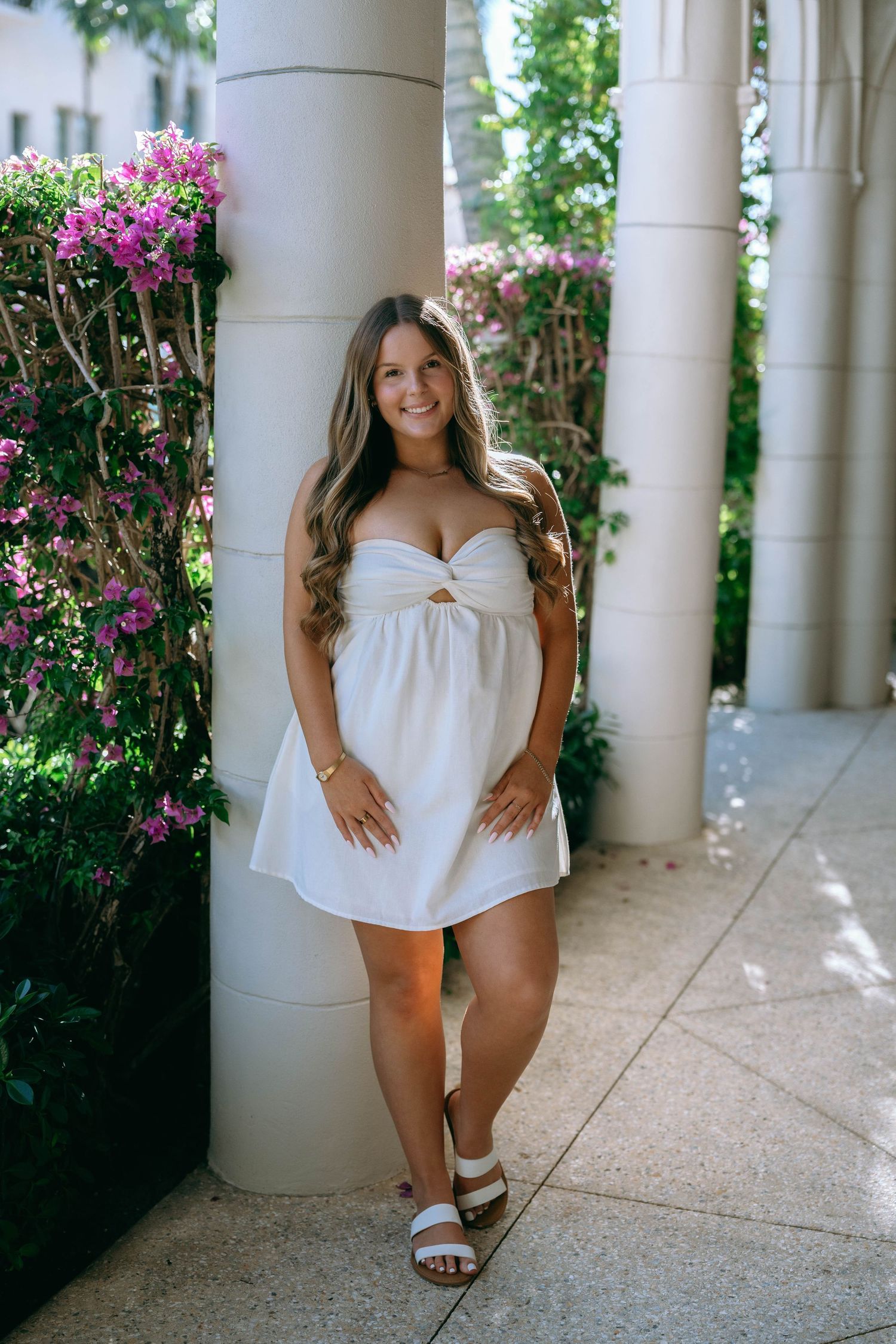 Young woman in white mini dress poses by floral columns in an elegant outdoor setting.