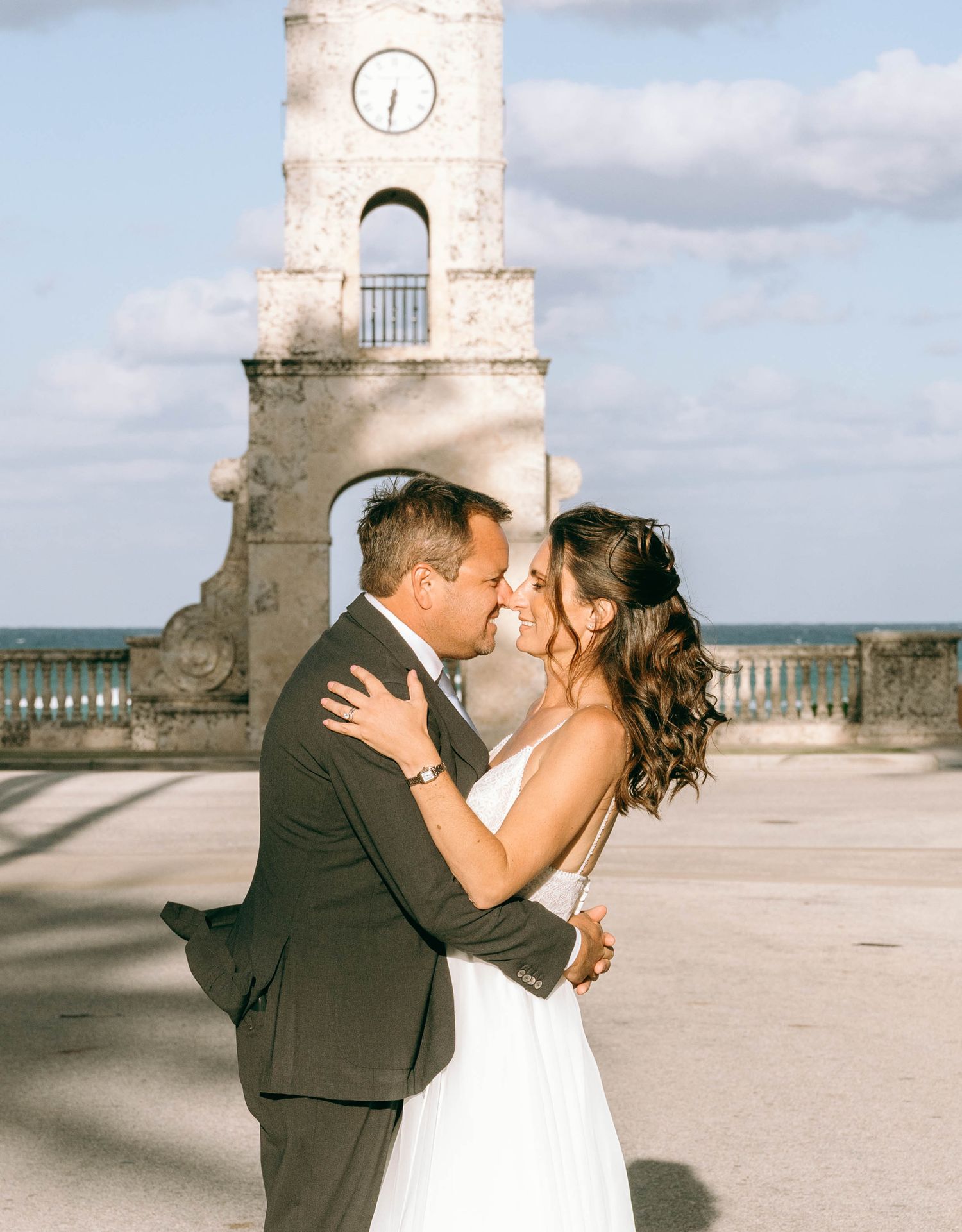 Bride and groom sharing romantic moment in front of historic clock tower near ocean waterfront.