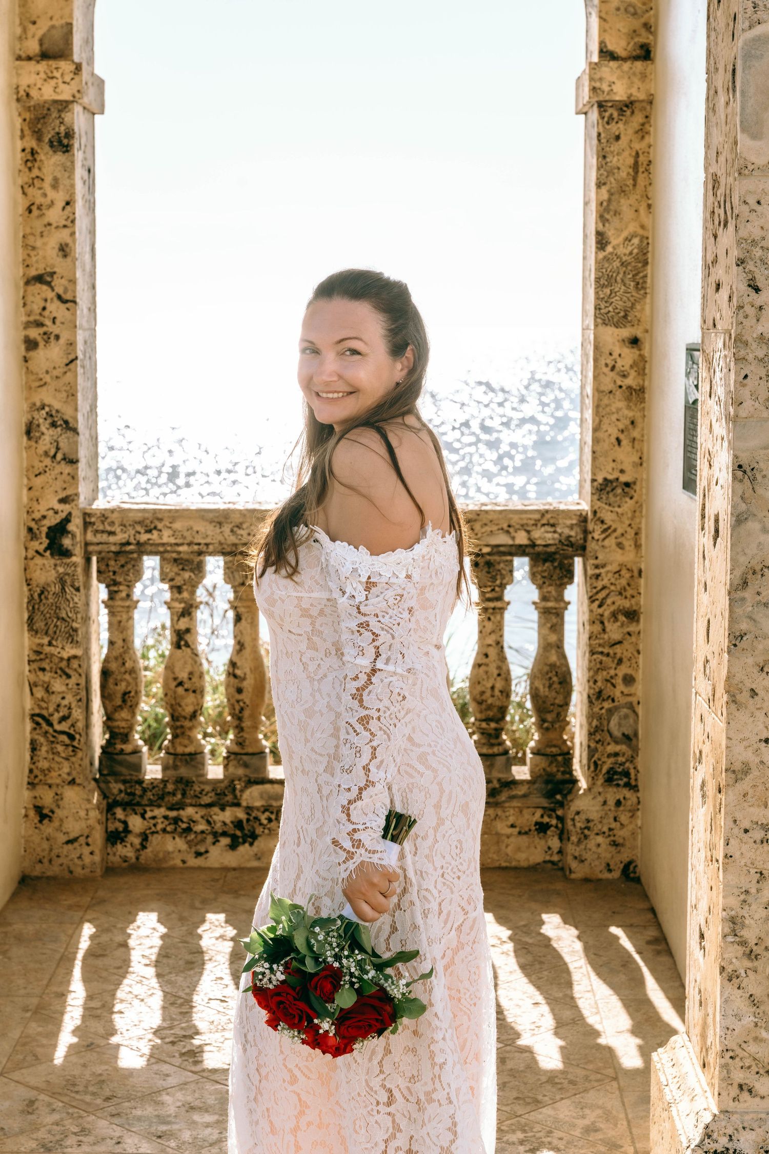 Smiling bride in off-shoulder lace gown holding red rose bouquet on sunlit stone terrace.