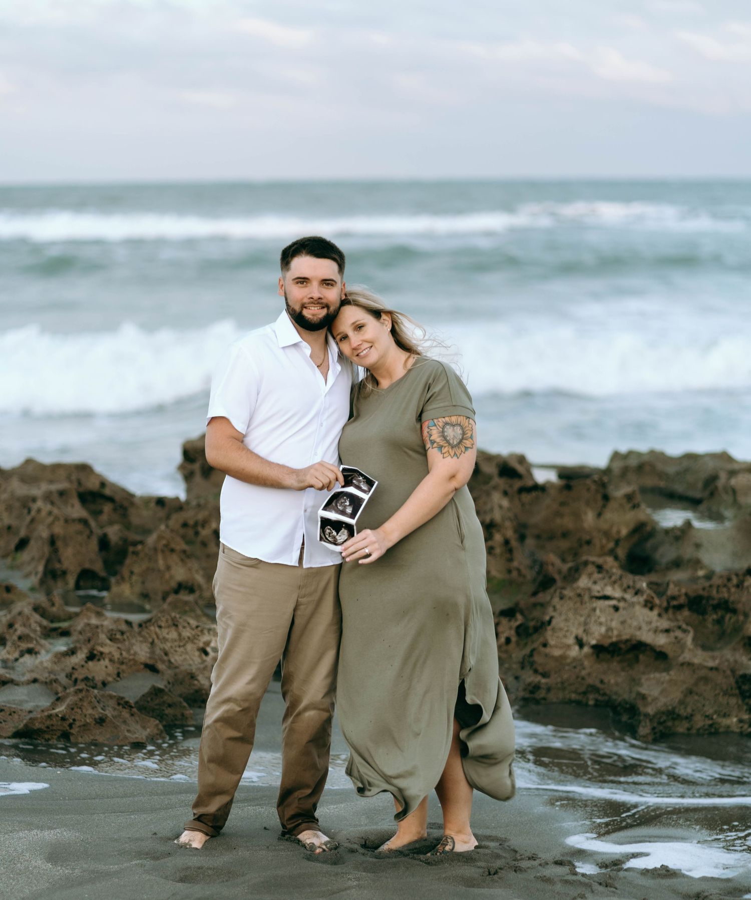 Romantic couple poses together on rocky beach during sunset maternity photoshoot.