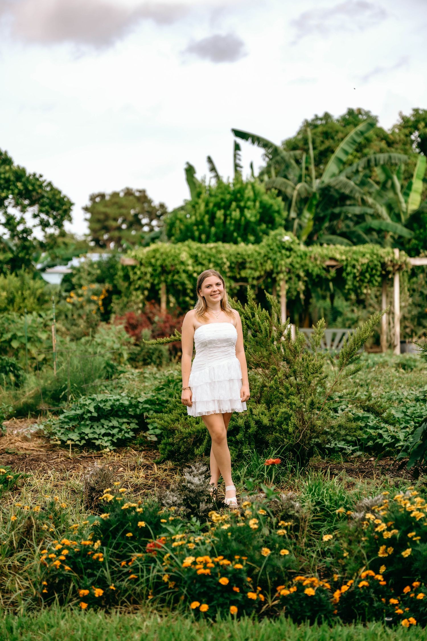 Young woman in white dress stands among colorful marigolds in a lush botanical garden.