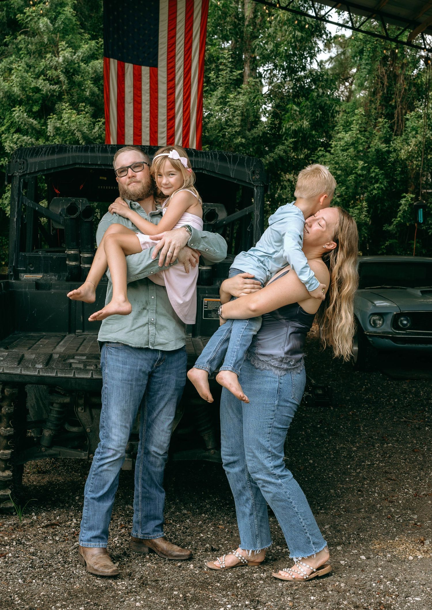 Family of four posing outdoors with American flag backdrop, parents holding children, wearing jeans and casual attire.