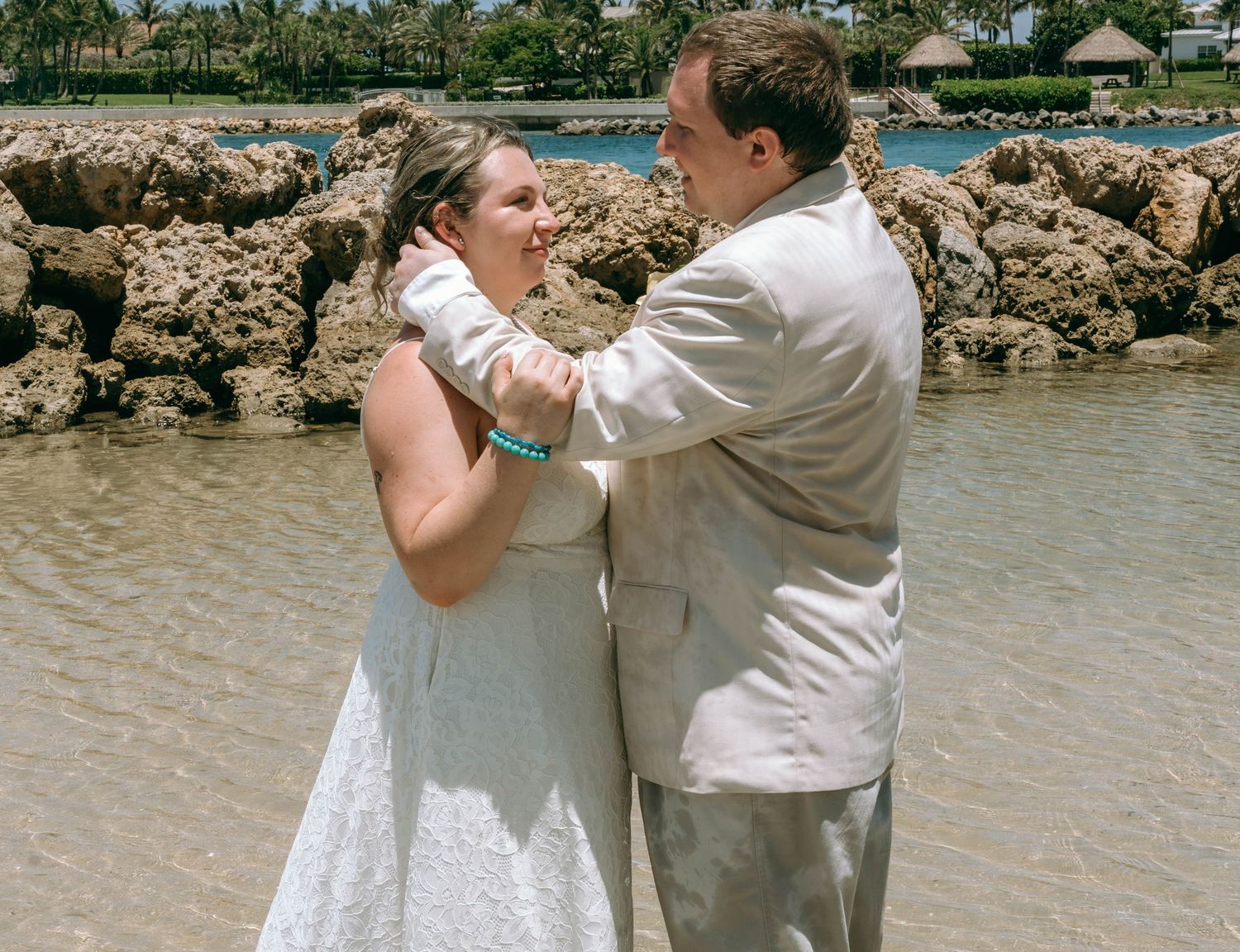 Bride and groom share a romantic moment by rocky shoreline, with bride in white dress and groom in light gray suit.