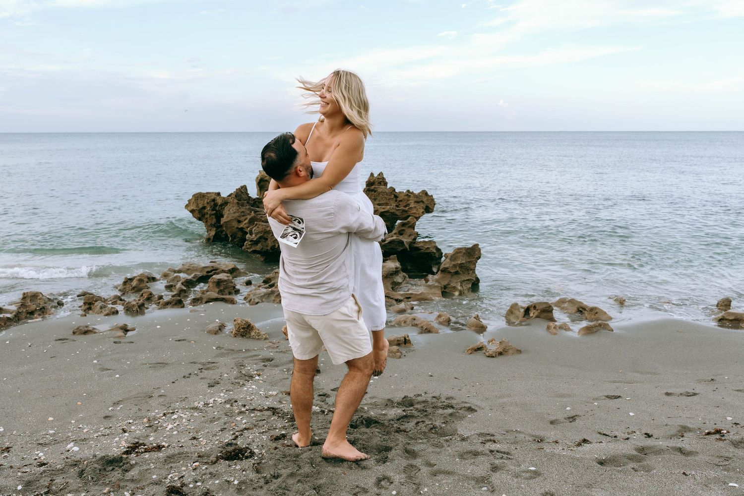 Couple embracing on a rocky beach shore, man lifting woman in white dress with ocean waves in background.