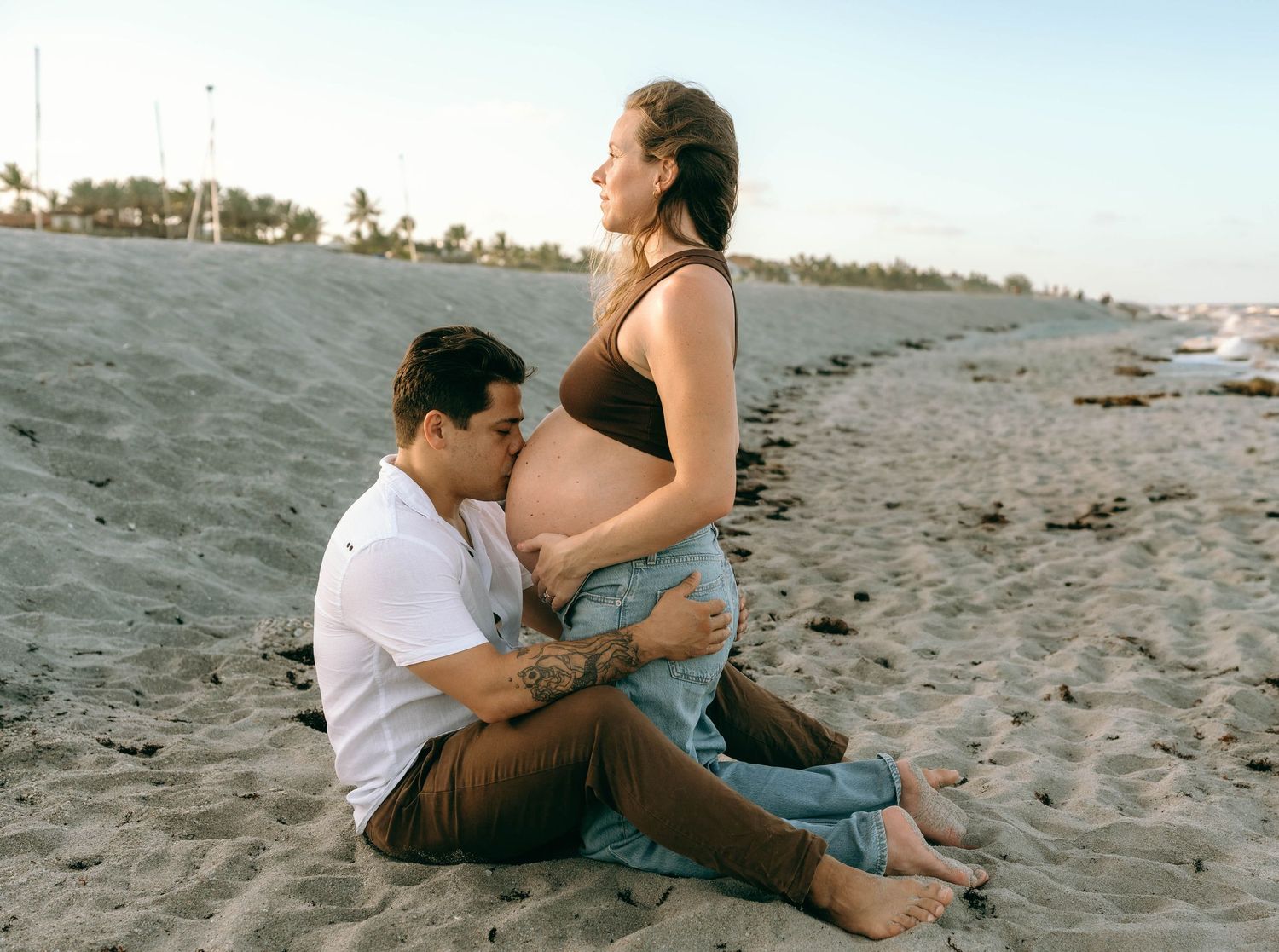 Couple sharing a tender maternity beach moment, man kissing pregnant belly while sitting on sandy shore at sunset.