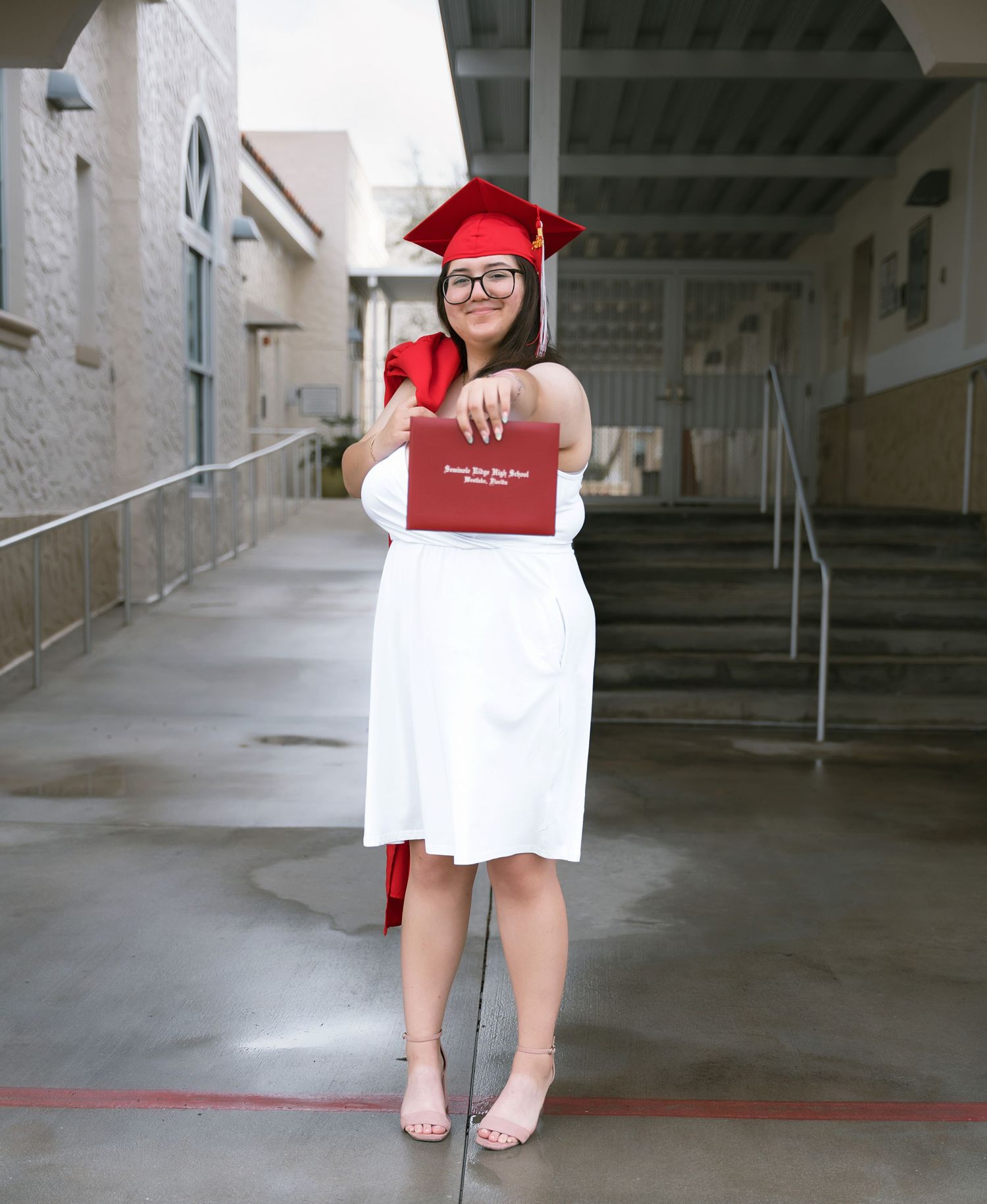 A graduate in white dress holds diploma while posing in academic regalia.